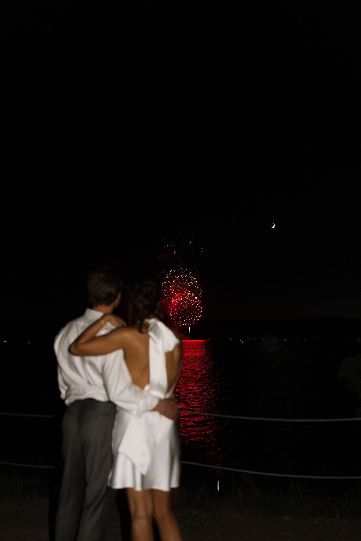 A couple embraces while watching red fireworks reflect off dark water at night.