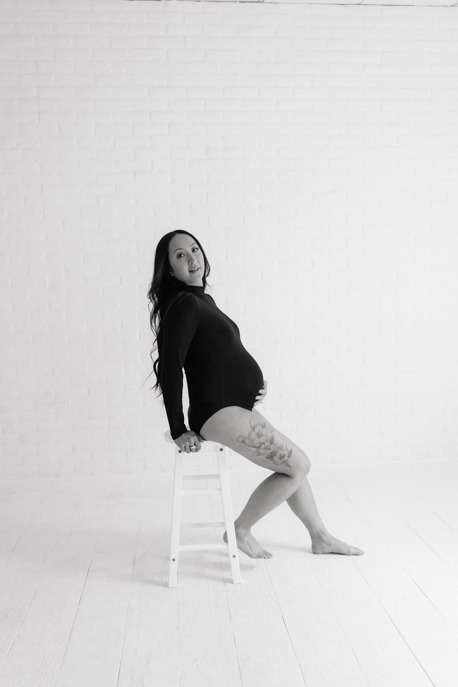 Black and white maternity photo of a woman in a bodysuit sitting on a white stool, showing her baby bump.