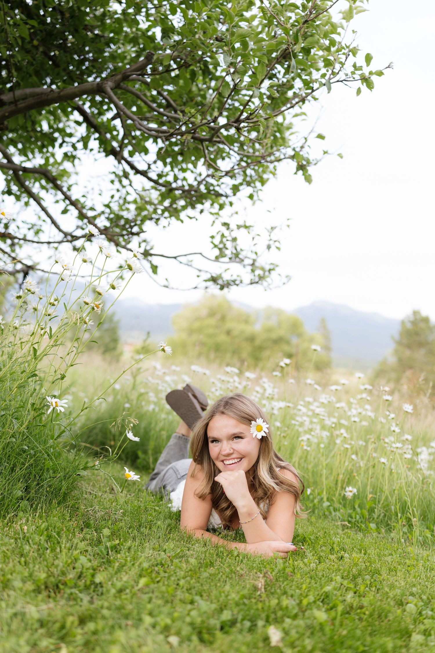 Young woman lying on green grass under a tree with a daisy in her hair, smiling in a wildflower meadow.