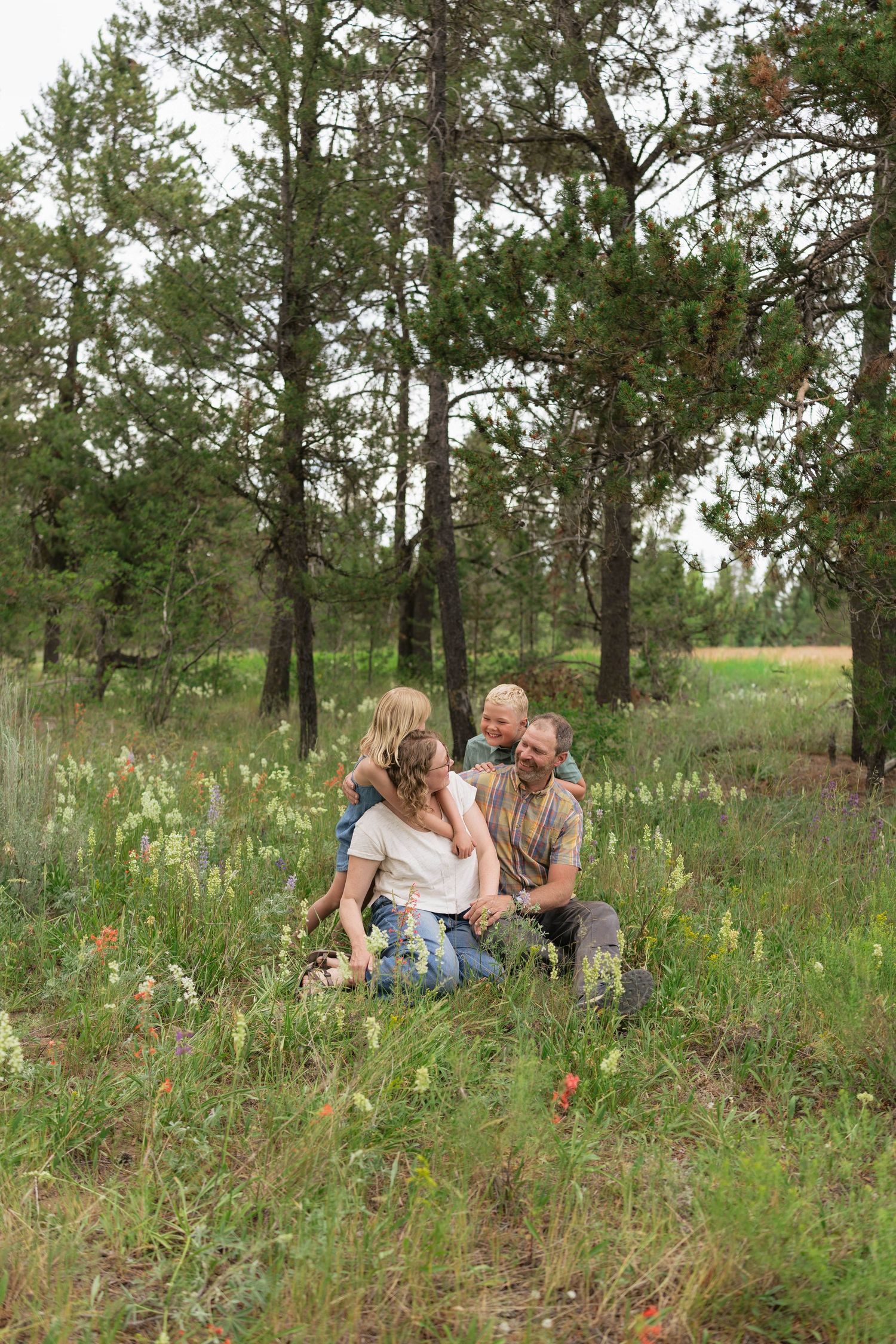 Family of four sitting together in a wildflower meadow surrounded by tall pine trees, sharing a joyful moment outdoors.