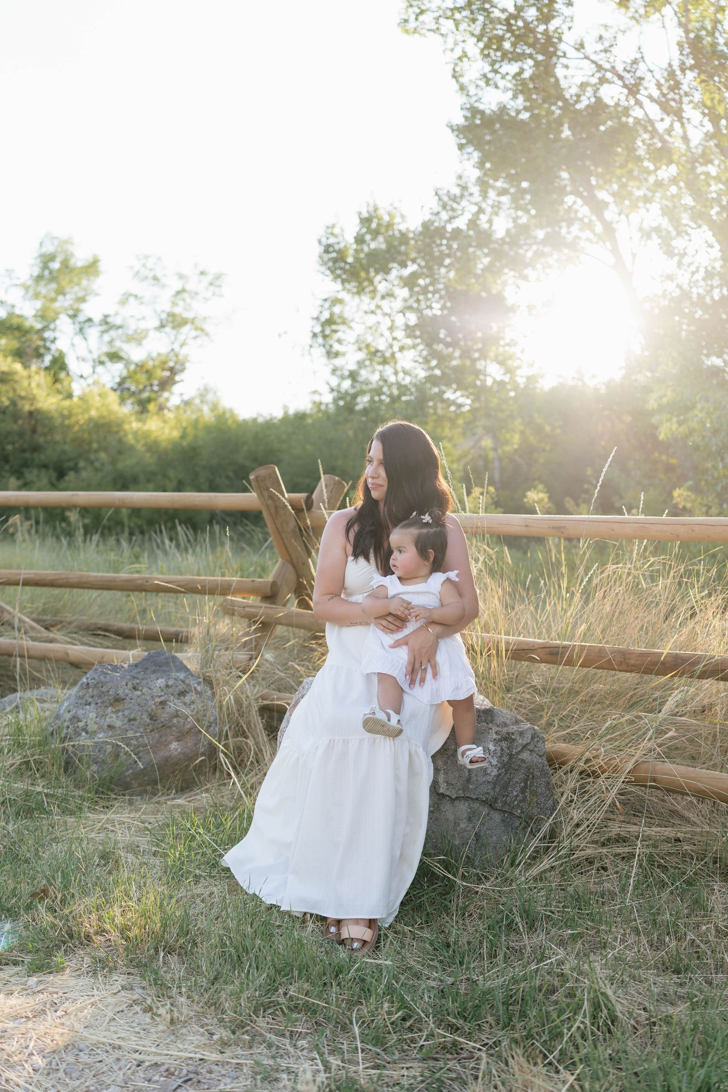 Mother in white dress holding toddler girl outdoors near rustic fence with golden sunlight through trees.