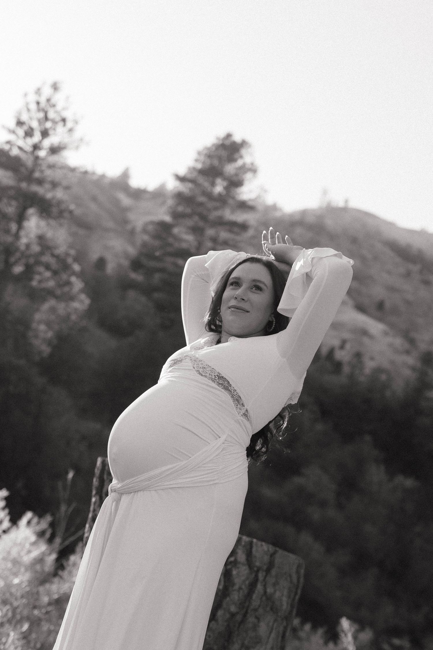 Pregnant woman in white gown and crown poses outdoors with arms raised, black and white maternity photo in nature.
