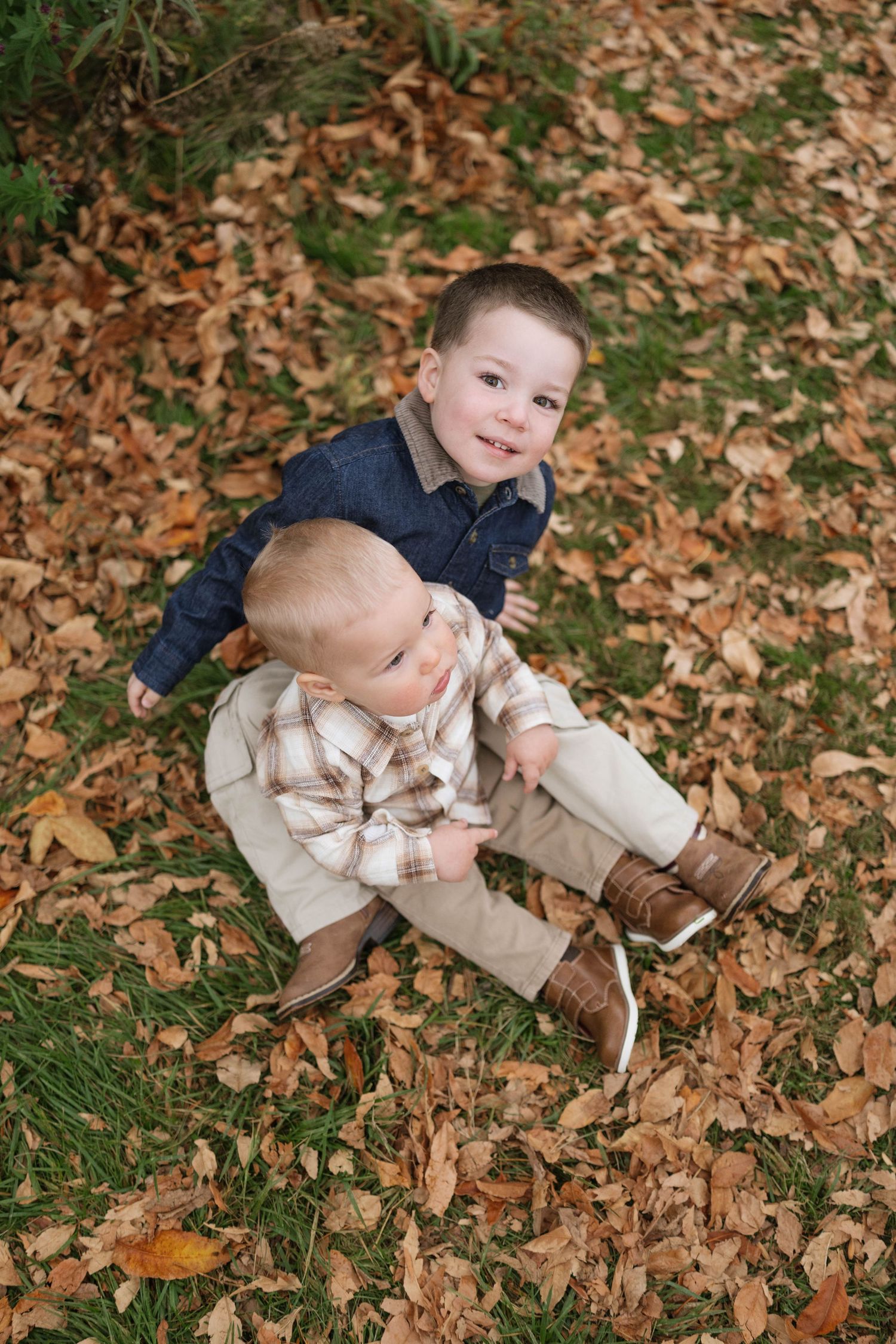 Two young boys in plaid and denim shirts sit together on grass covered with fallen autumn leaves during a fall family photo session.