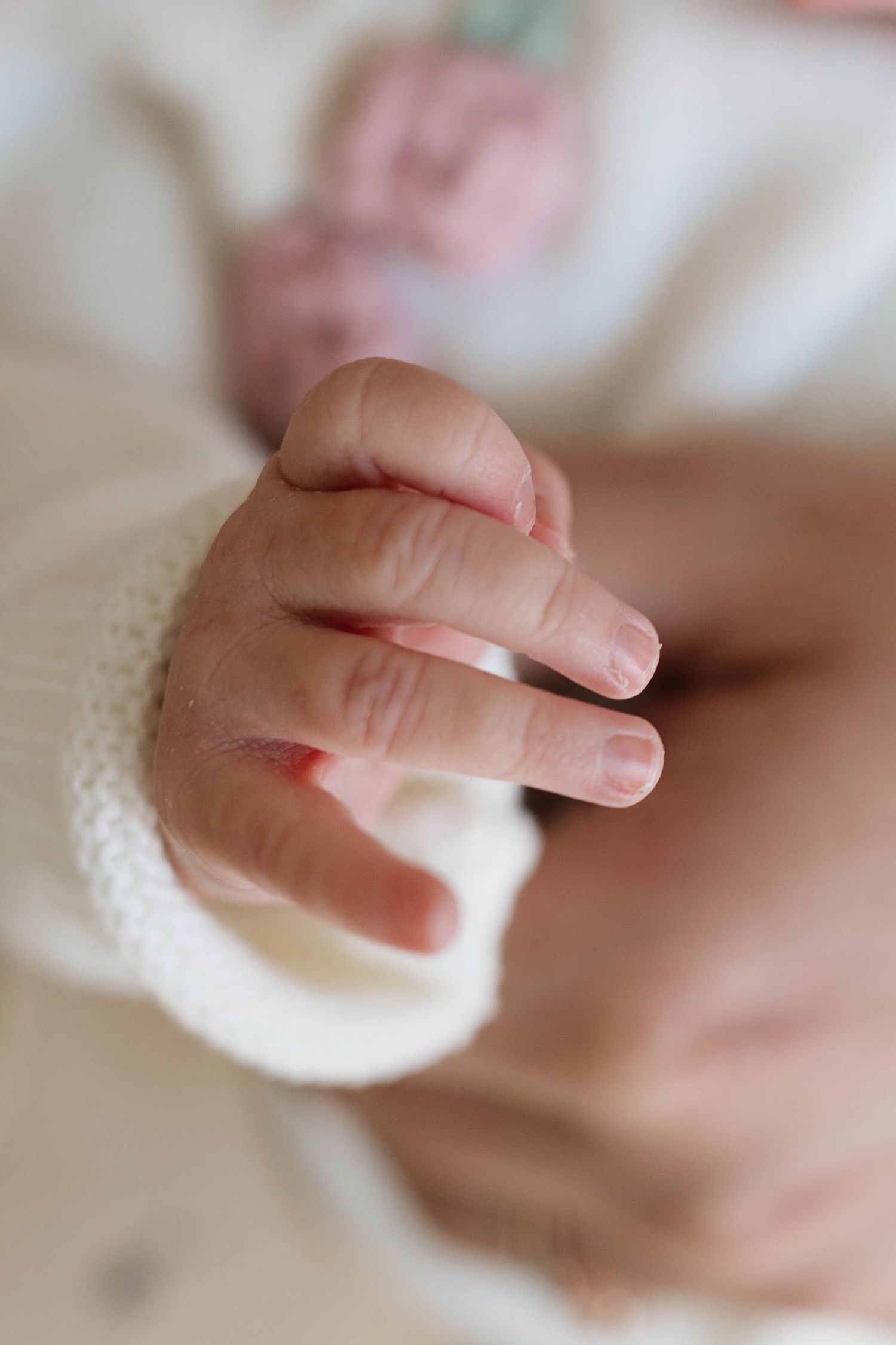 Close-up of a newborn baby's tiny curled hand with delicate fingers, wrapped in a soft white knit sleeve with a blurred pink detail.