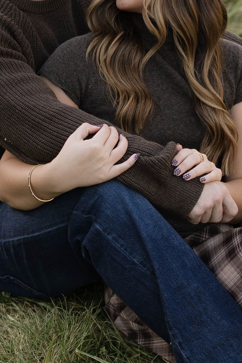 Couple sitting close on the grass in cozy sweaters and jeans, with arms wrapped around each other in a warm, romantic outdoor moment.