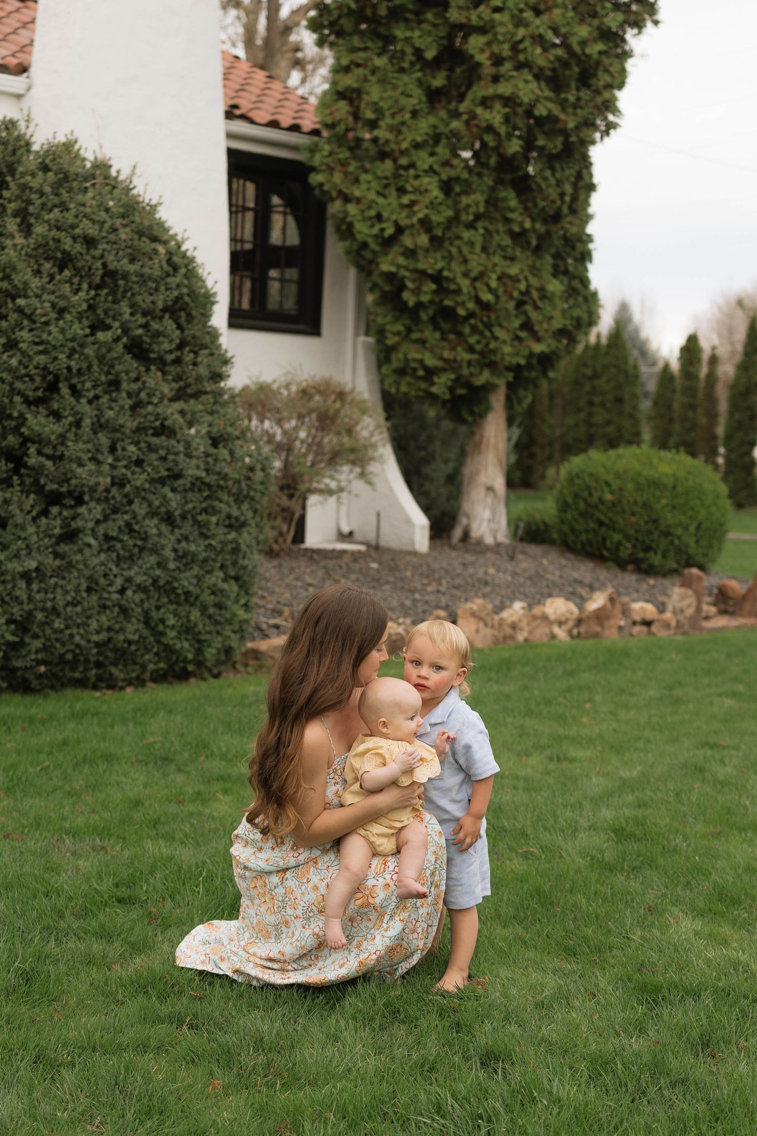 A tender family moment captured on a lush green lawn beside a house with climbing vines and manicured hedges.