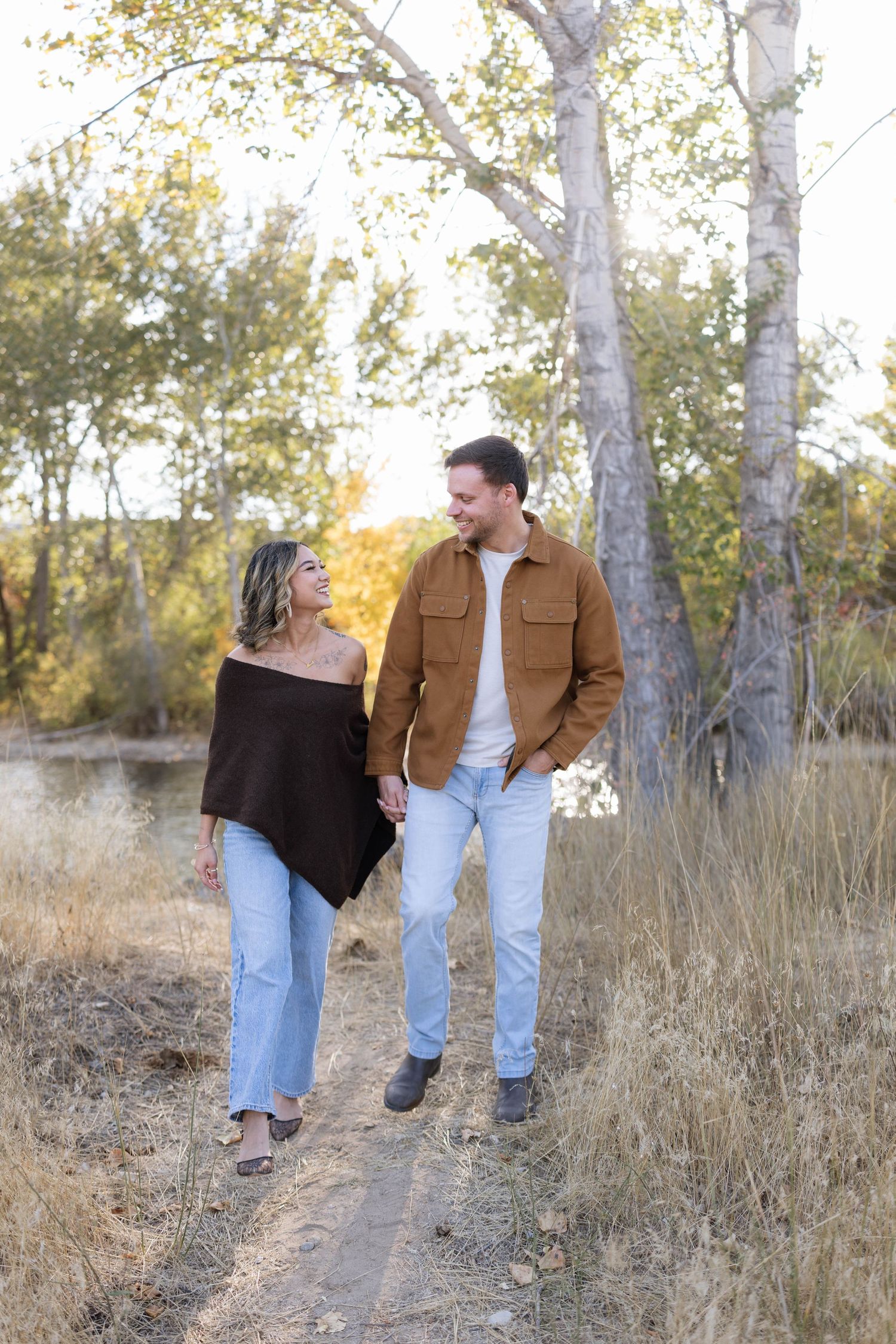 Smiling couple in matching light-wash jeans walk hand in hand along a dirt path through golden autumn grass and tall trees.