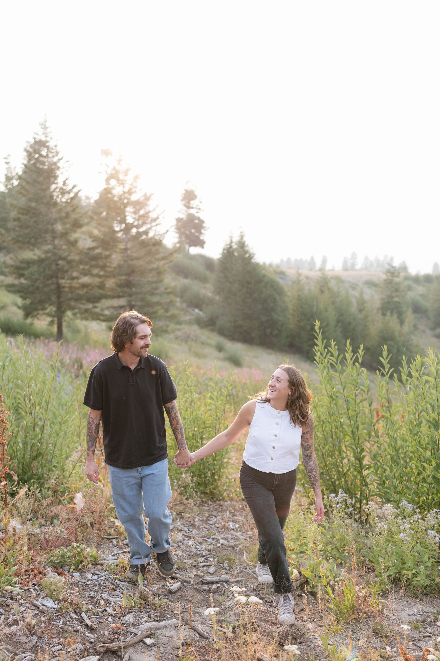 Smiling couple holding hands and walking along a dirt trail through wildflowers and pine trees in warm golden hour light.