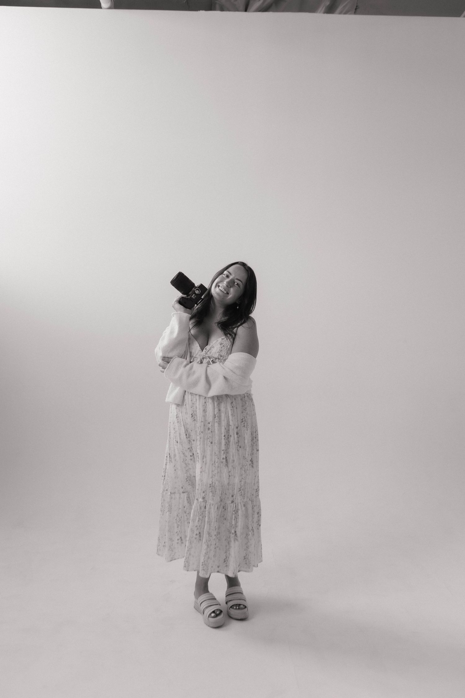 Black and white photo of a woman in a floral dress holding a camera and looking up in a bright studio.