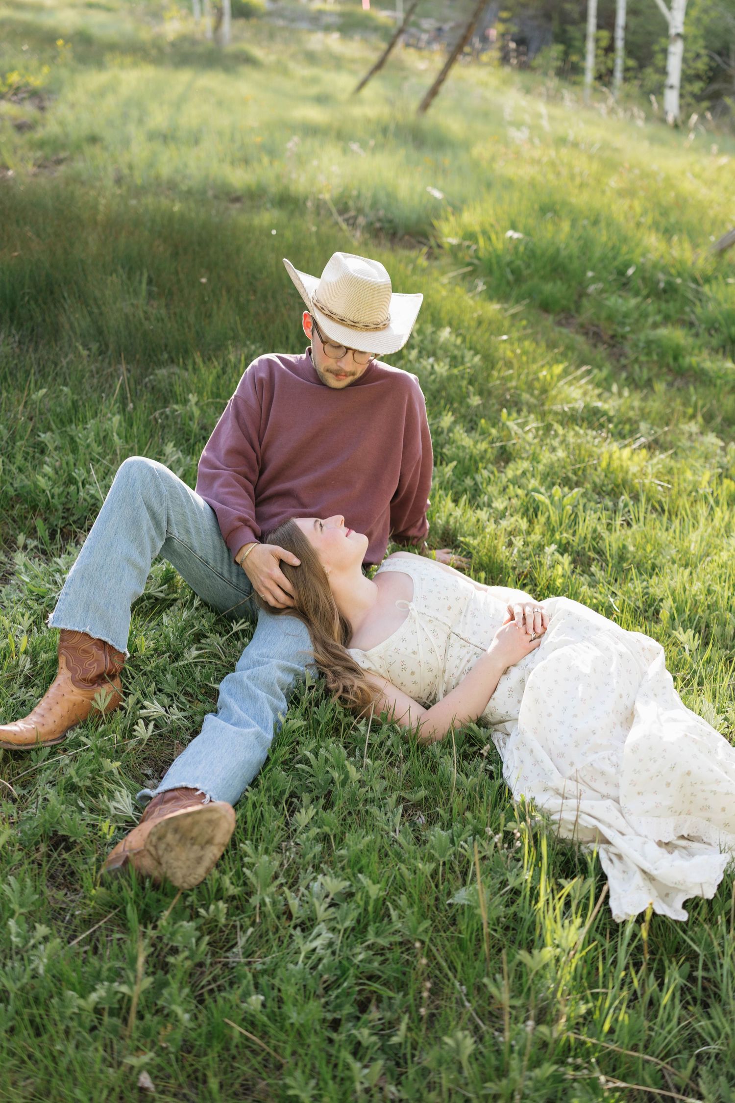 A couple relaxes on green grass, man in cowboy hat and boots sits while woman in white dress lays across his lap.