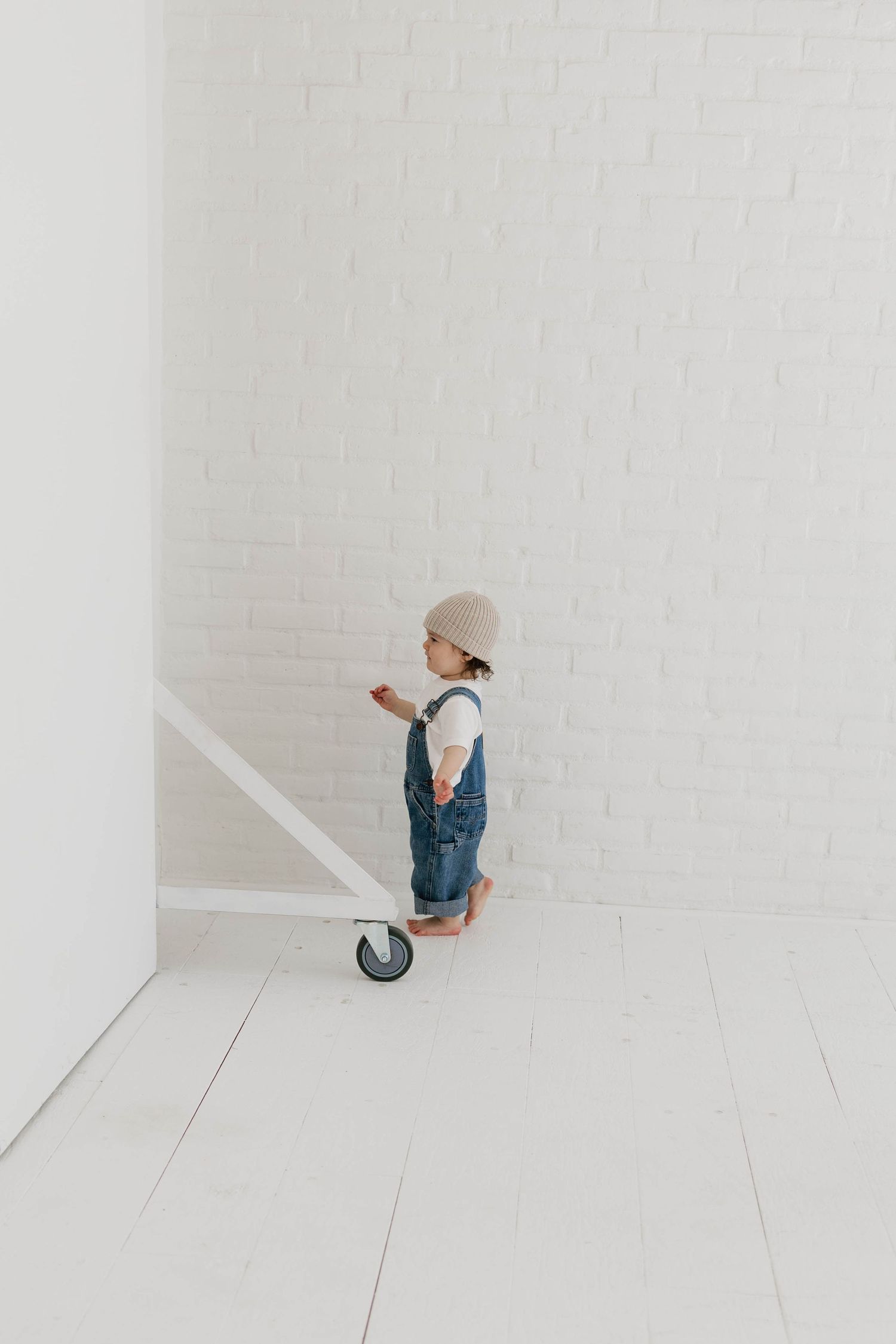 Toddler in denim overalls and beige hat stands barefoot in a minimalist white room with brick wall.