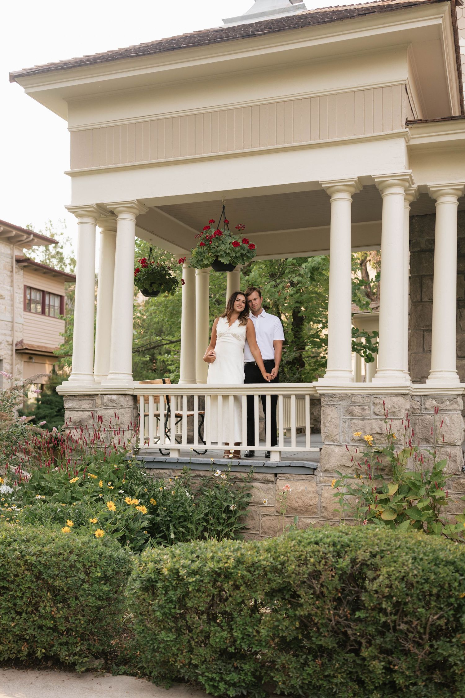 Couple in white dress and black pants posing on elegant columned porch surrounded by lush gardens.