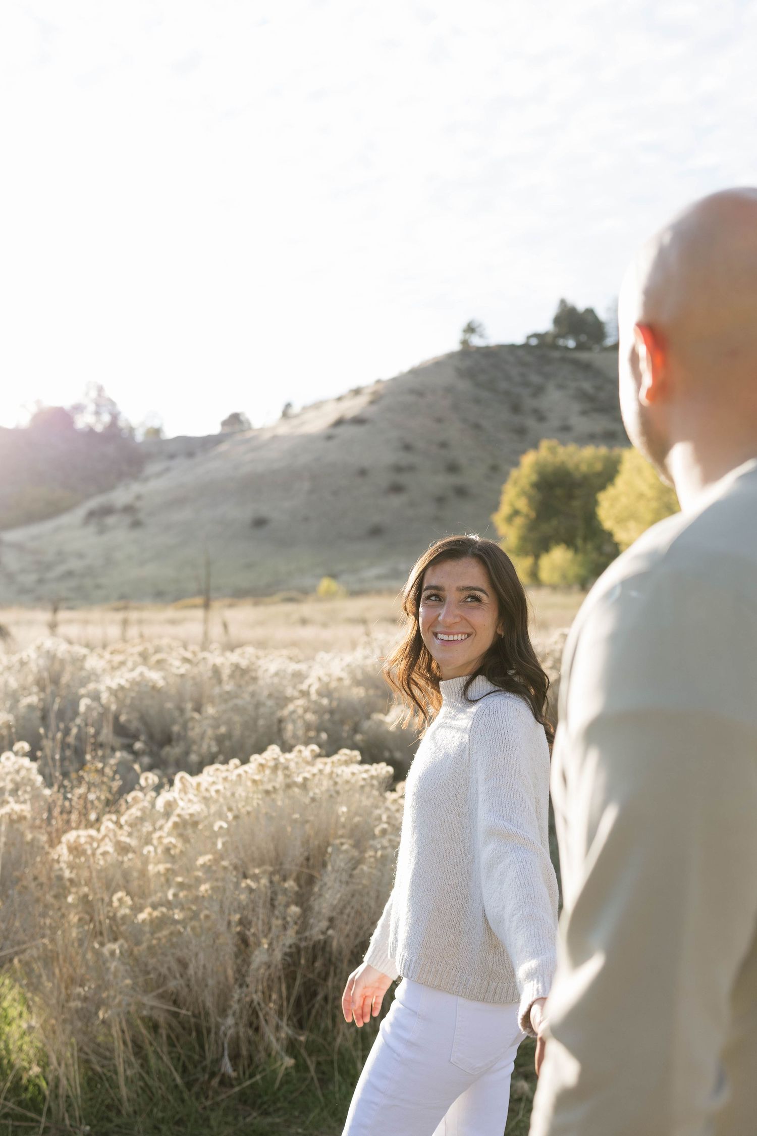 Couple poses in golden field at sunset with mountains in background during romantic photo session.