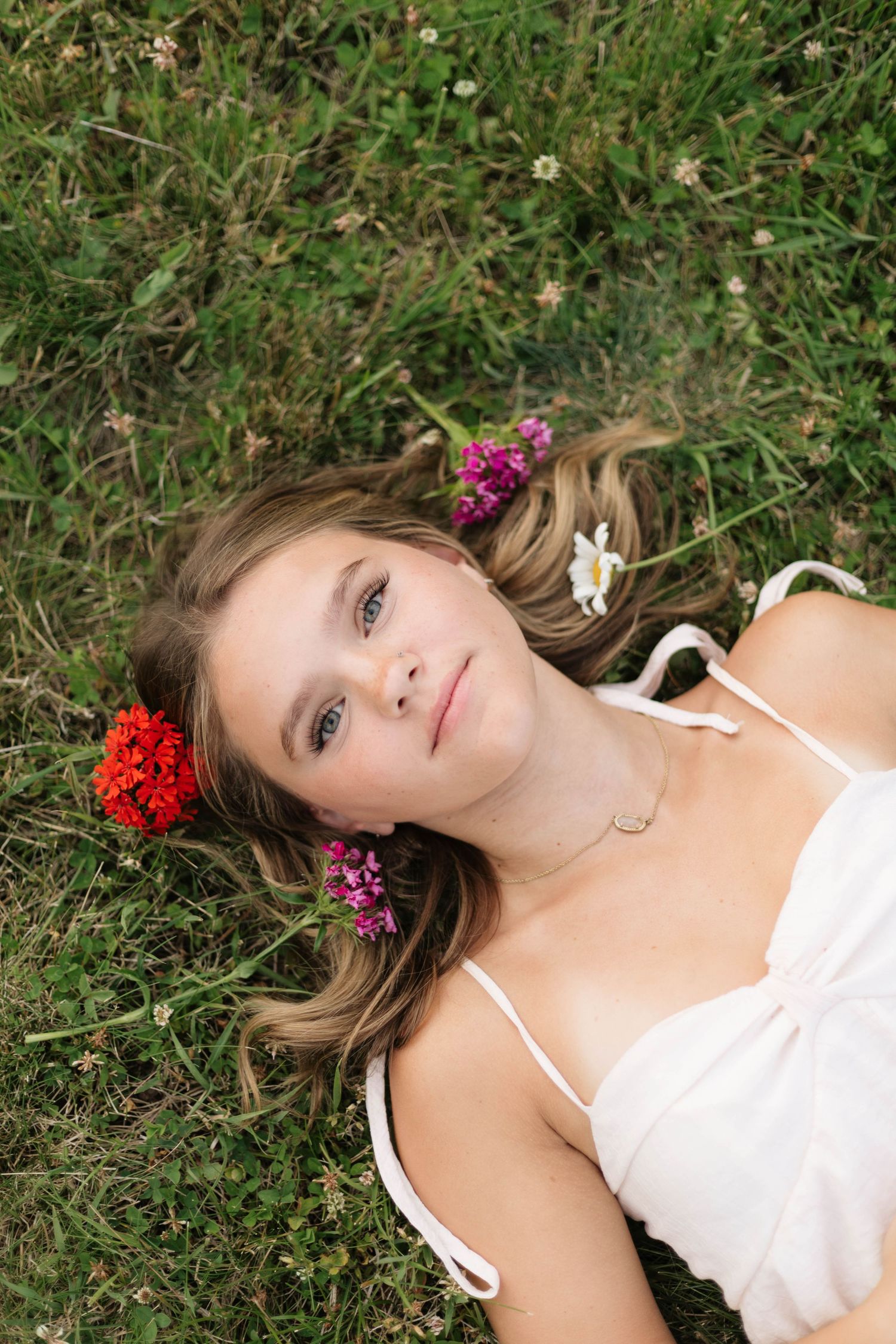 Young woman lying in green grass with colorful wildflowers in her hair, wearing a white dress, looking up peacefully.