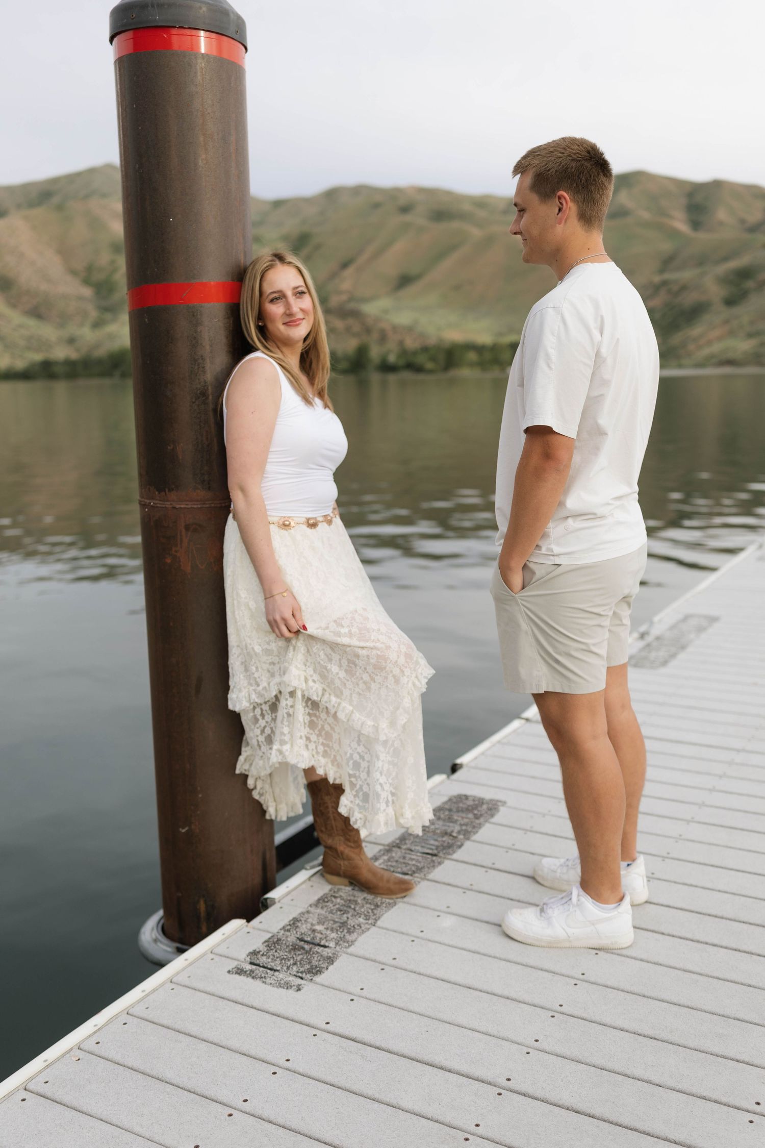 Young couple in white outfits sharing a moment on a dock by a serene lake with scenic mountain backdrop.