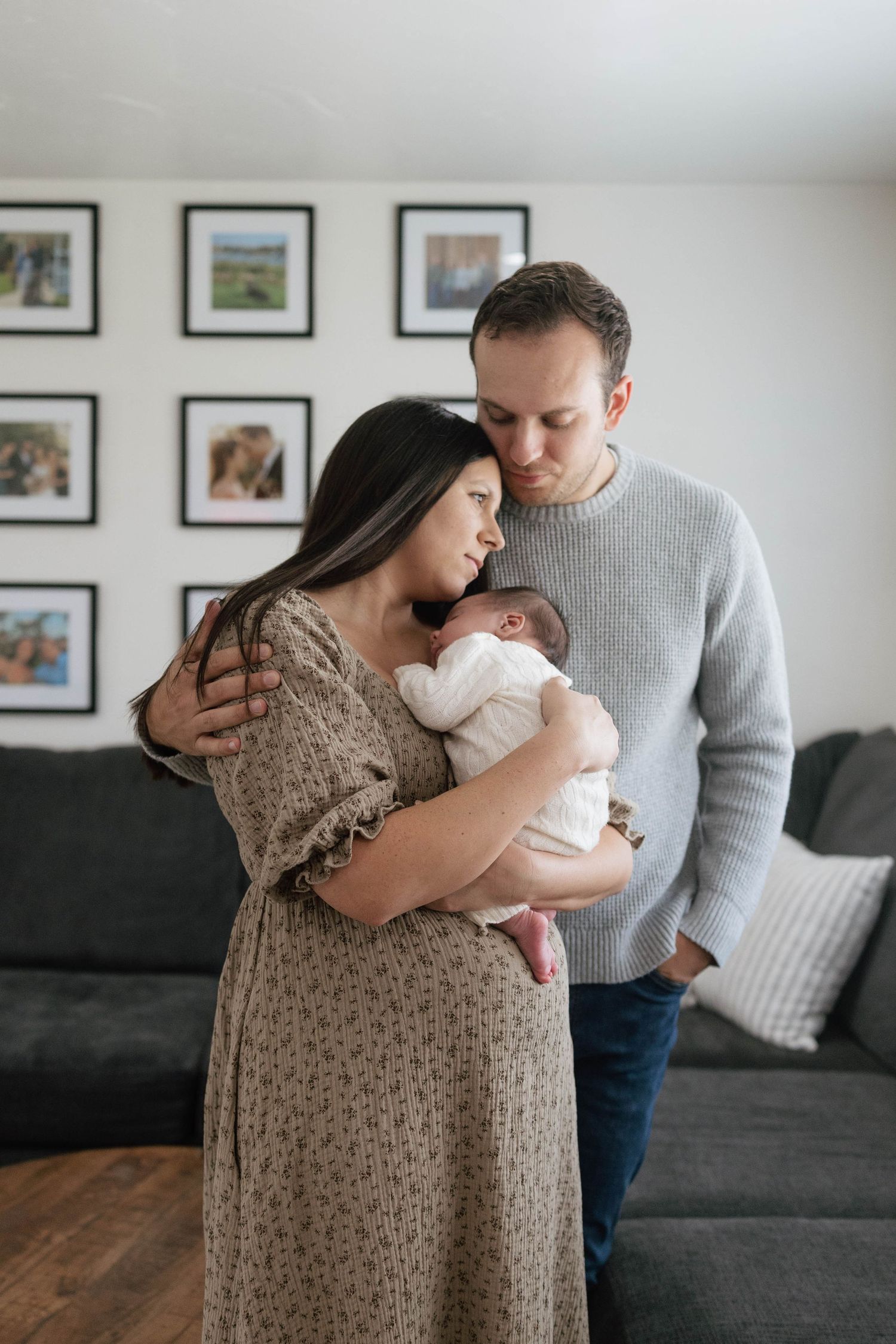 Parents share tender moments with their newborn baby in a cozy living room with framed photos on the wall.