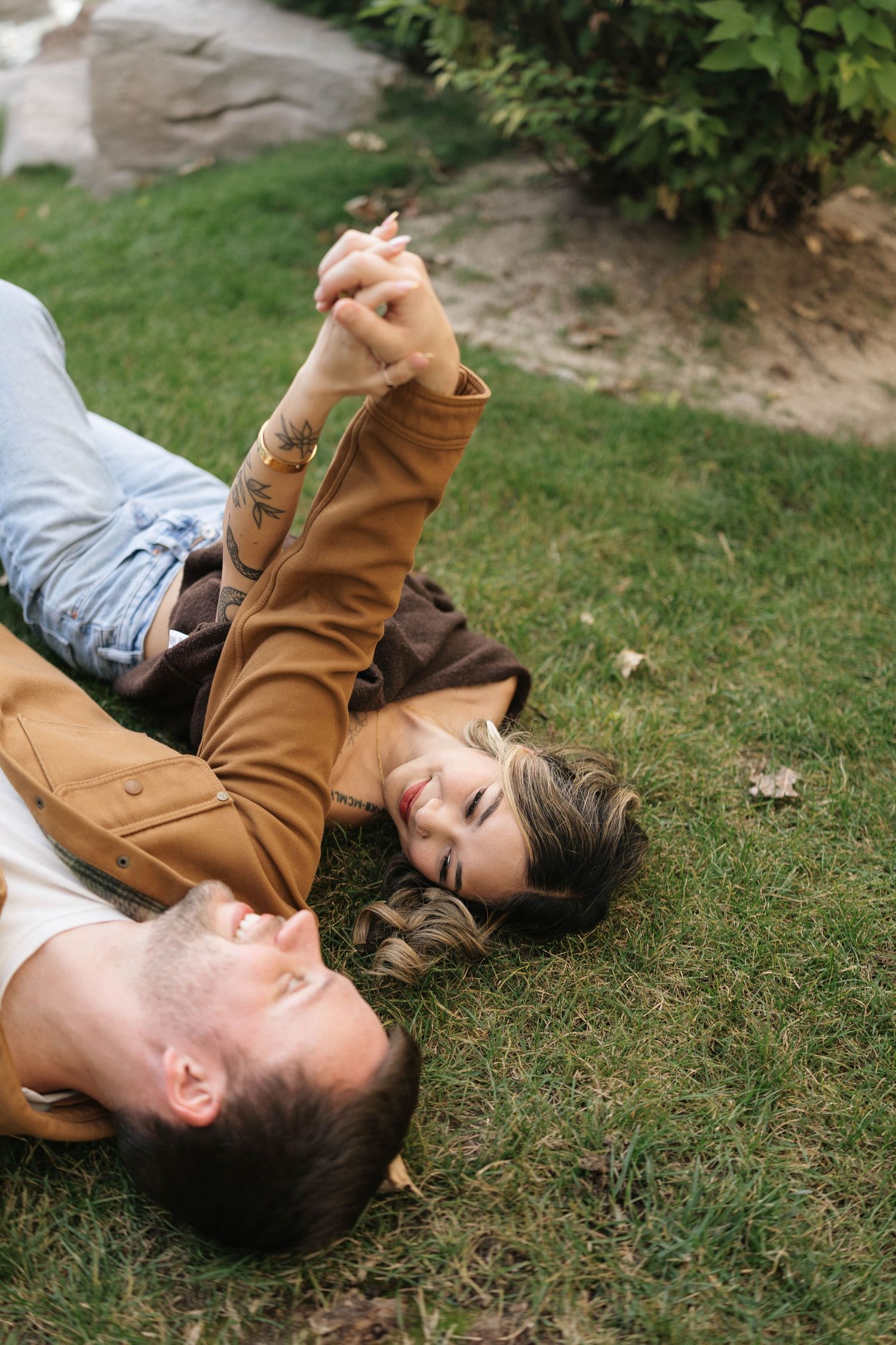 Smiling couple lying on green grass holding hands in the air, wearing casual tan jackets, enjoying a relaxed romantic moment outdoors.