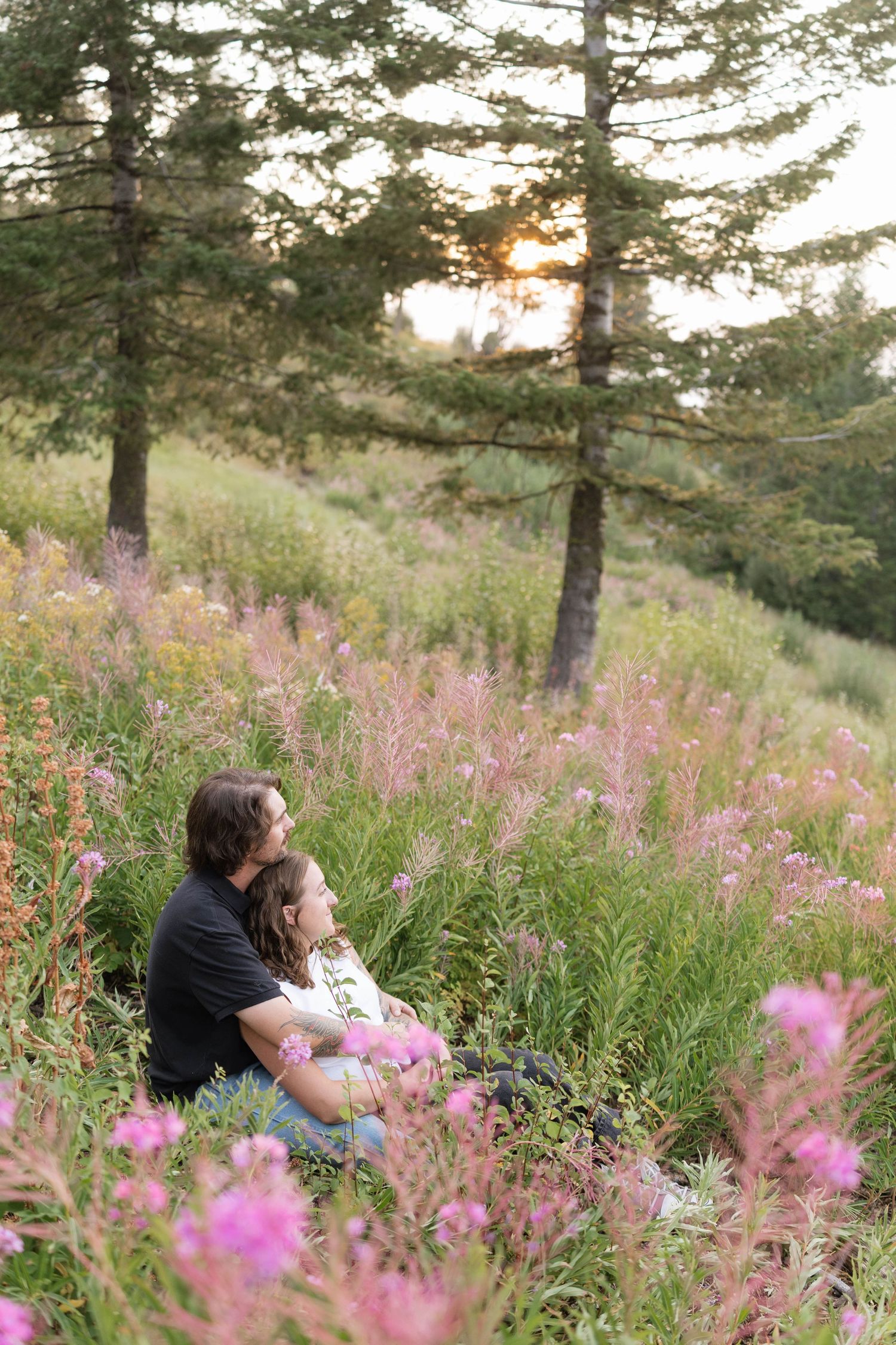Couple sitting close together in a meadow of pink fireweed wildflowers, watching the sunset glow through tall evergreen trees.