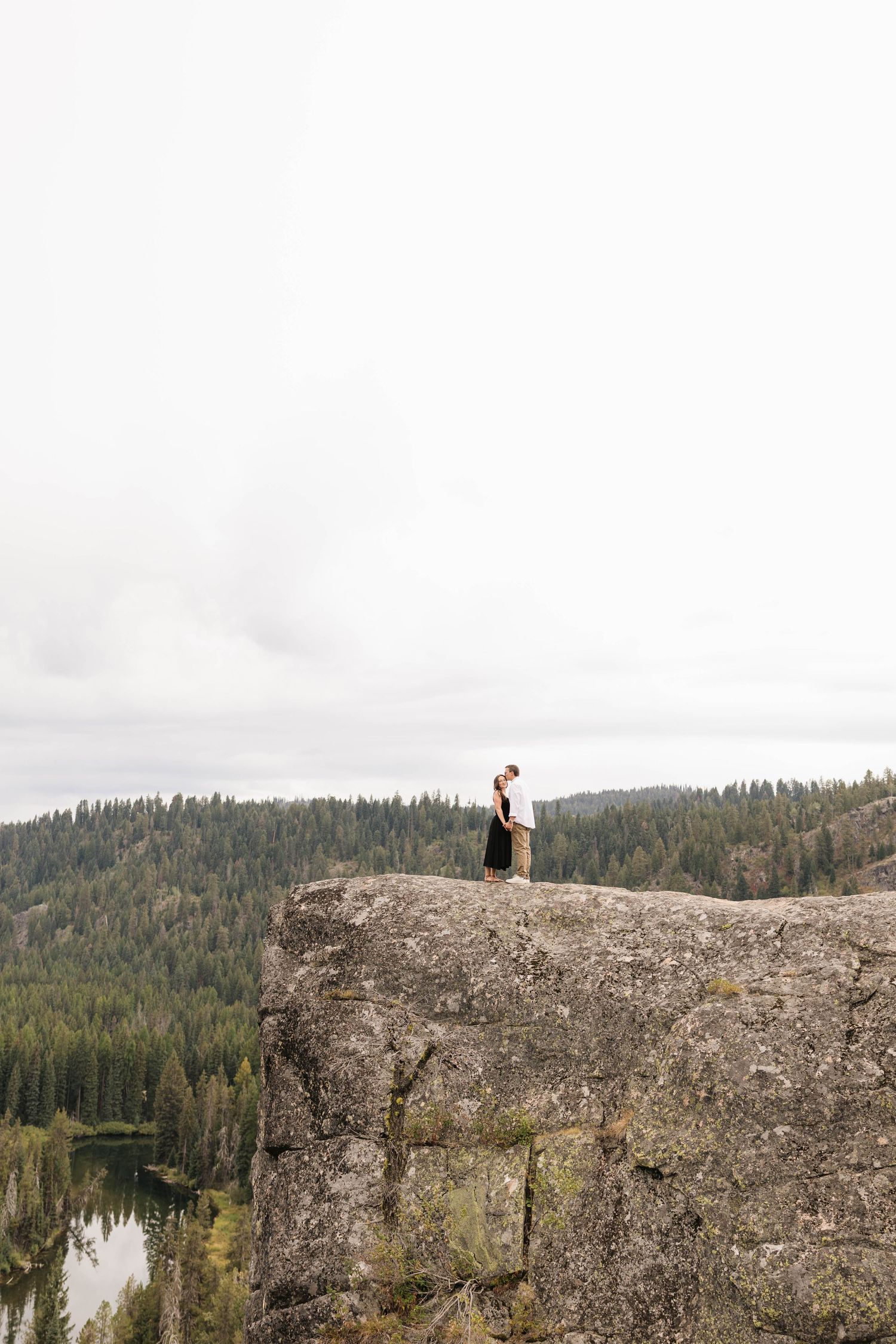 Couple standing on a rocky cliff edge overlooking a dense pine forest and lake on an overcast day.