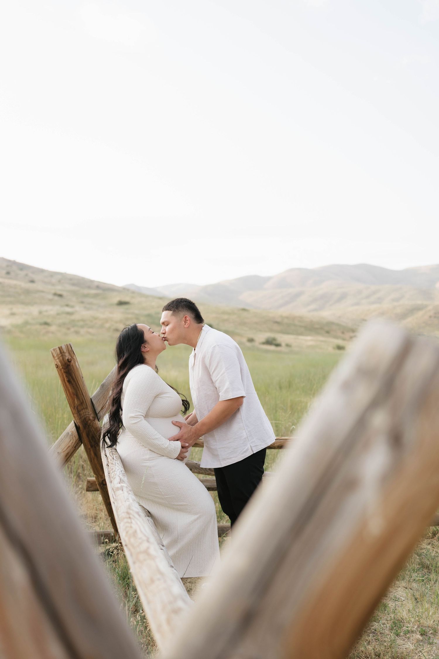 Pregnant couple sharing a kiss by a rustic wooden fence with scenic mountain meadow backdrop.