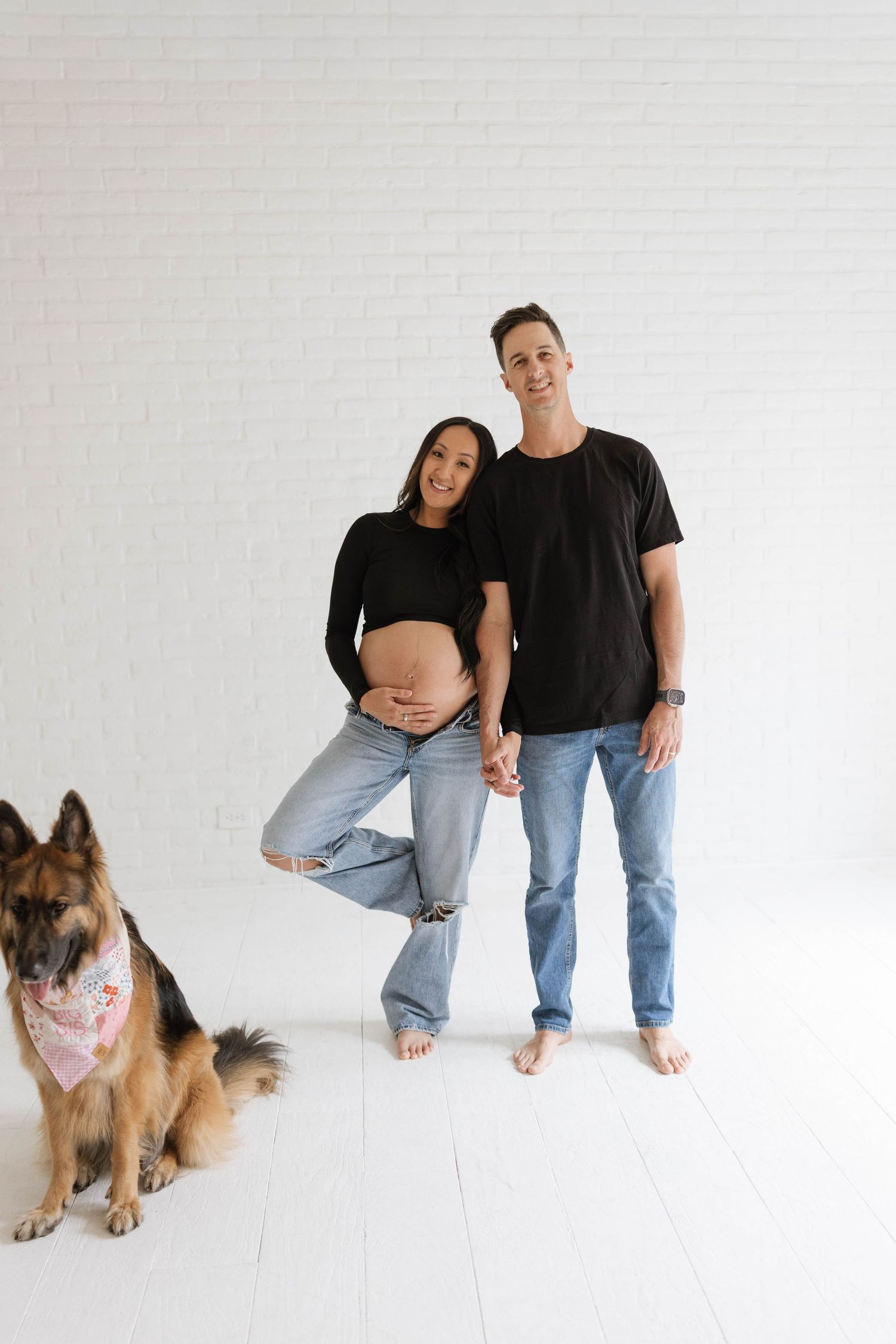 Pregnant couple in black tops and jeans posing with a German Shepherd dog wearing a pink bandana against white brick wall.
