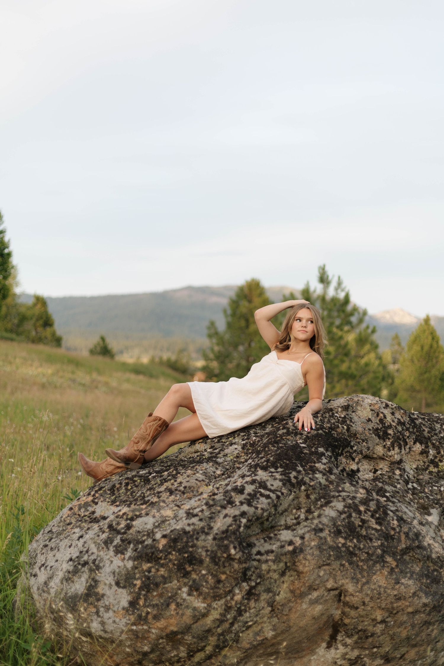 Young woman in white dress and cowboy boots reclines on a large boulder in a scenic mountain meadow.