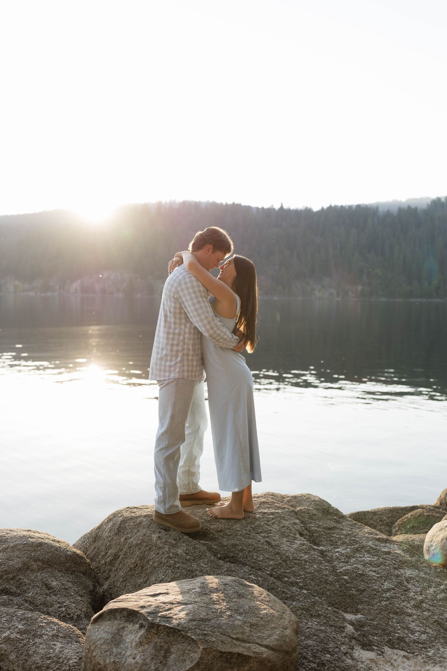 A couple shares a romantic moment on rocky lakeside cliffs at sunset, with serene water views in the background.