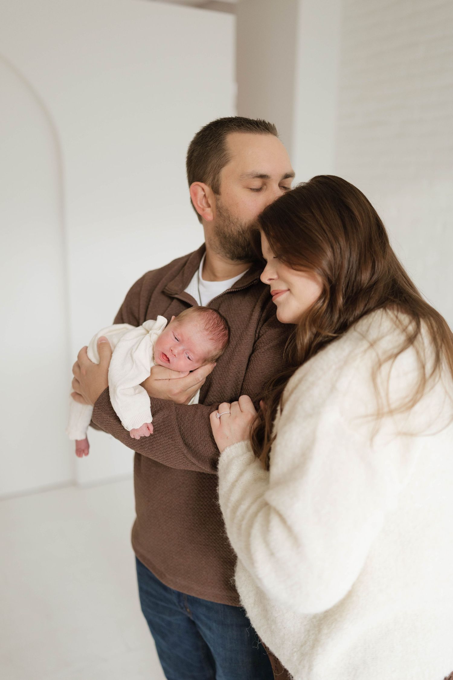 A couple shares an intimate moment while holding their newborn baby in a bright white room.