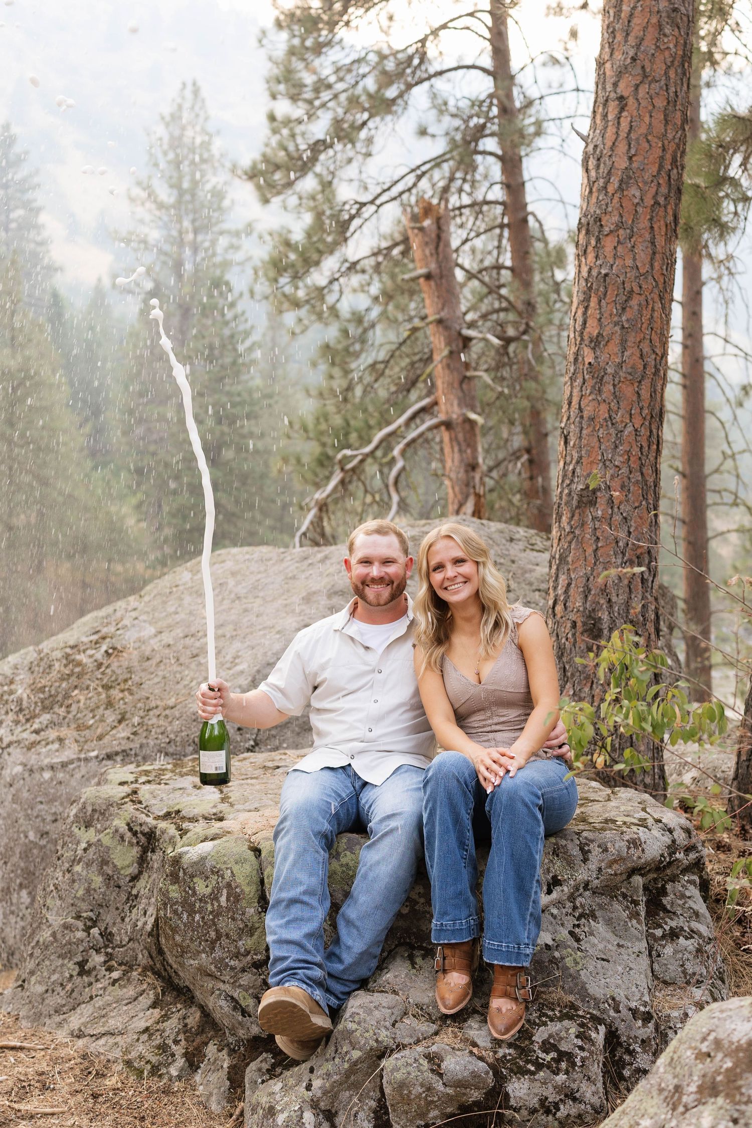 Couple sitting on rocks in a forest celebrating with champagne bottle popping, surrounded by pine trees and misty mountains.