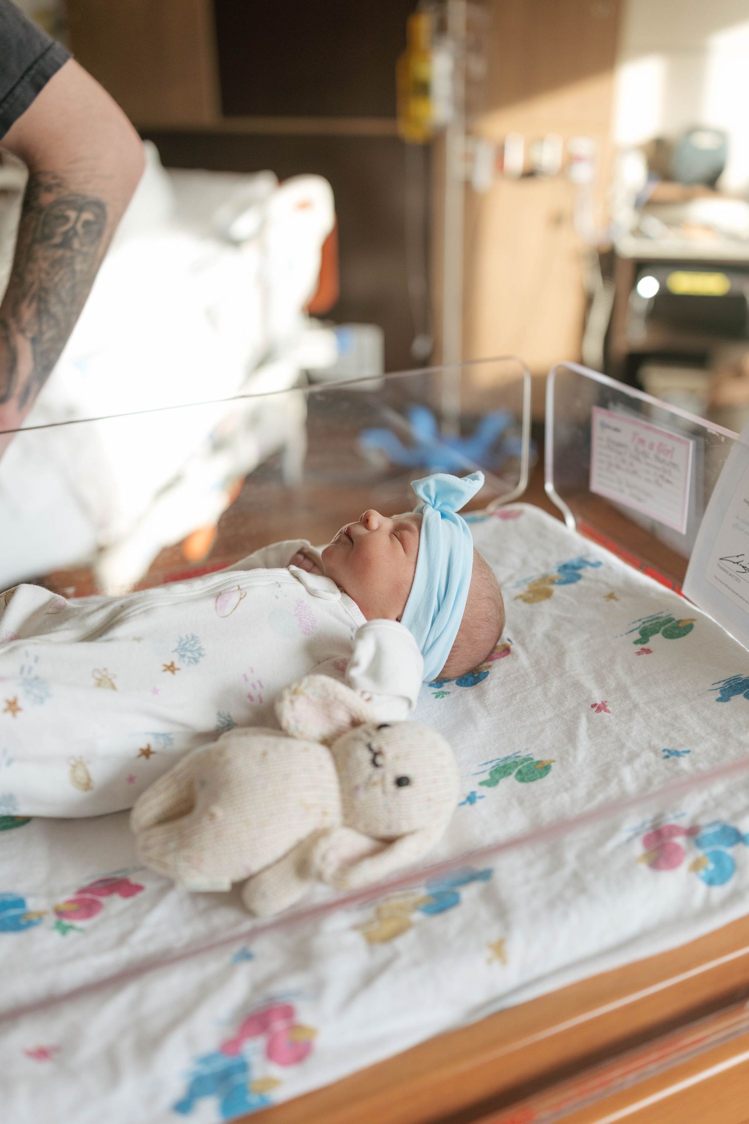 A premature baby sleeps peacefully in a hospital incubator with a medical mask and floral patterned blanket.