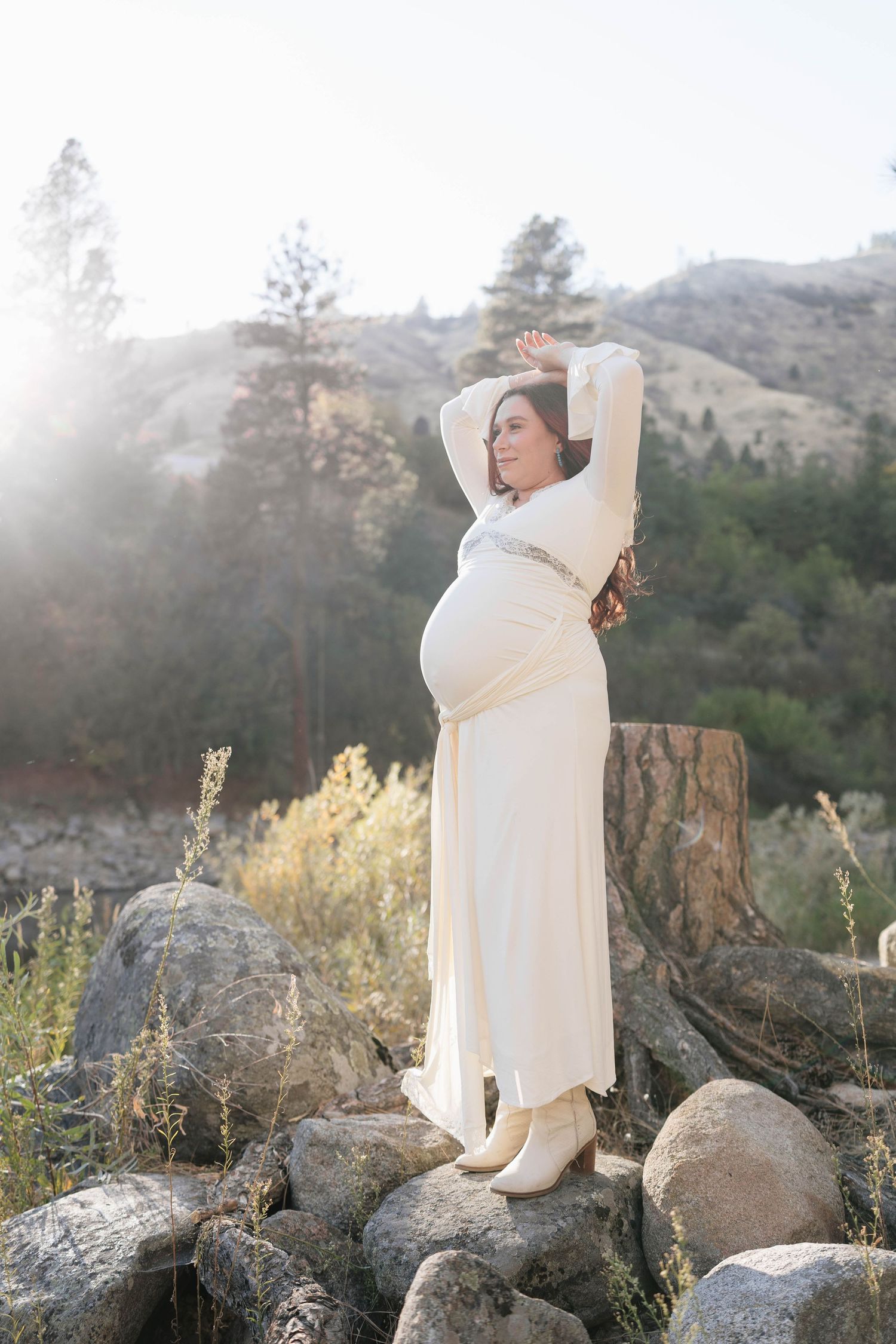 Pregnant woman in a cream flowing dress and boots poses with raised arms on rocks in a sunlit mountain landscape for maternity photos.