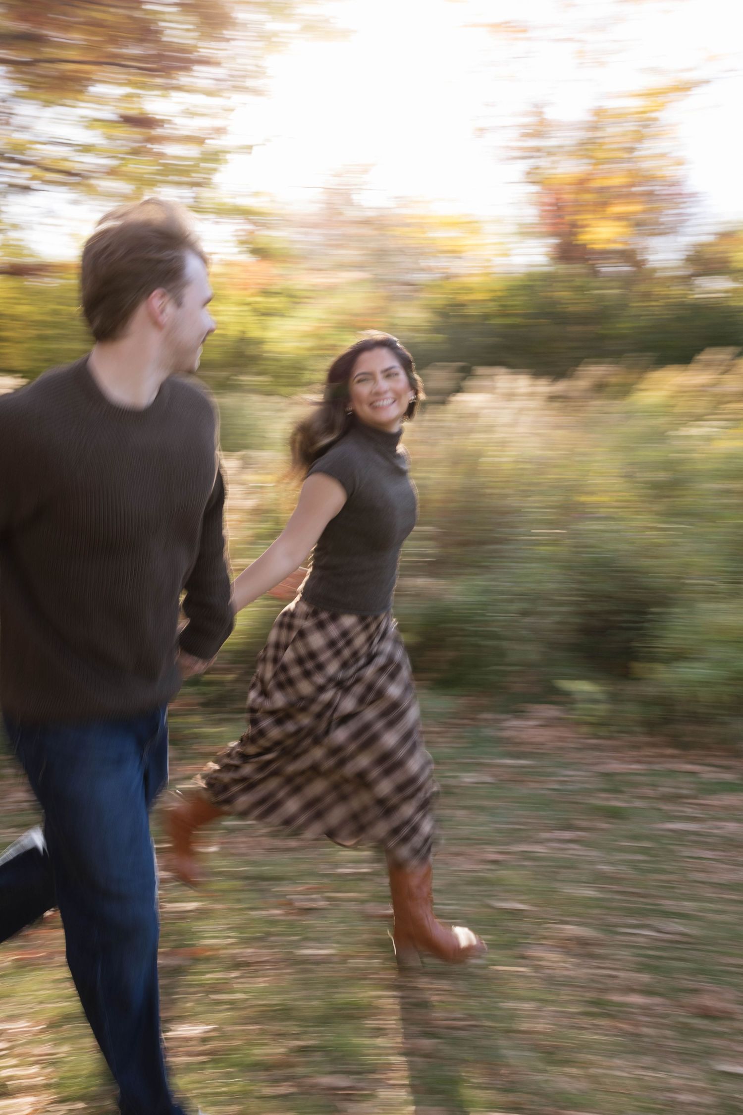 Smiling woman in a plaid skirt and brown boots runs hand in hand with a man through a sunlit autumn park in a motion-blurred photo.