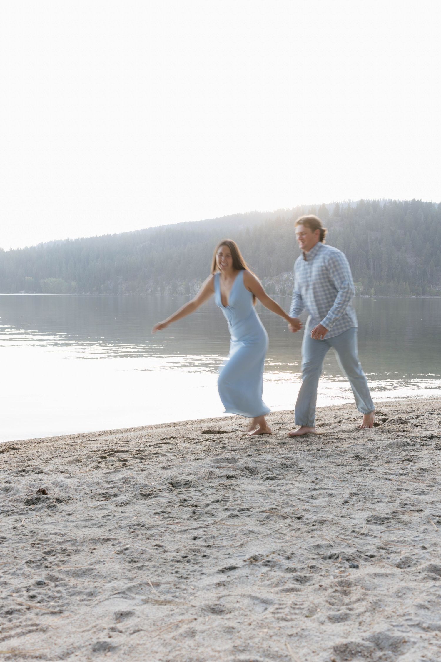 A couple in light blue outfits plays together on a sandy beach beside a serene lake with mountains in the distance.
