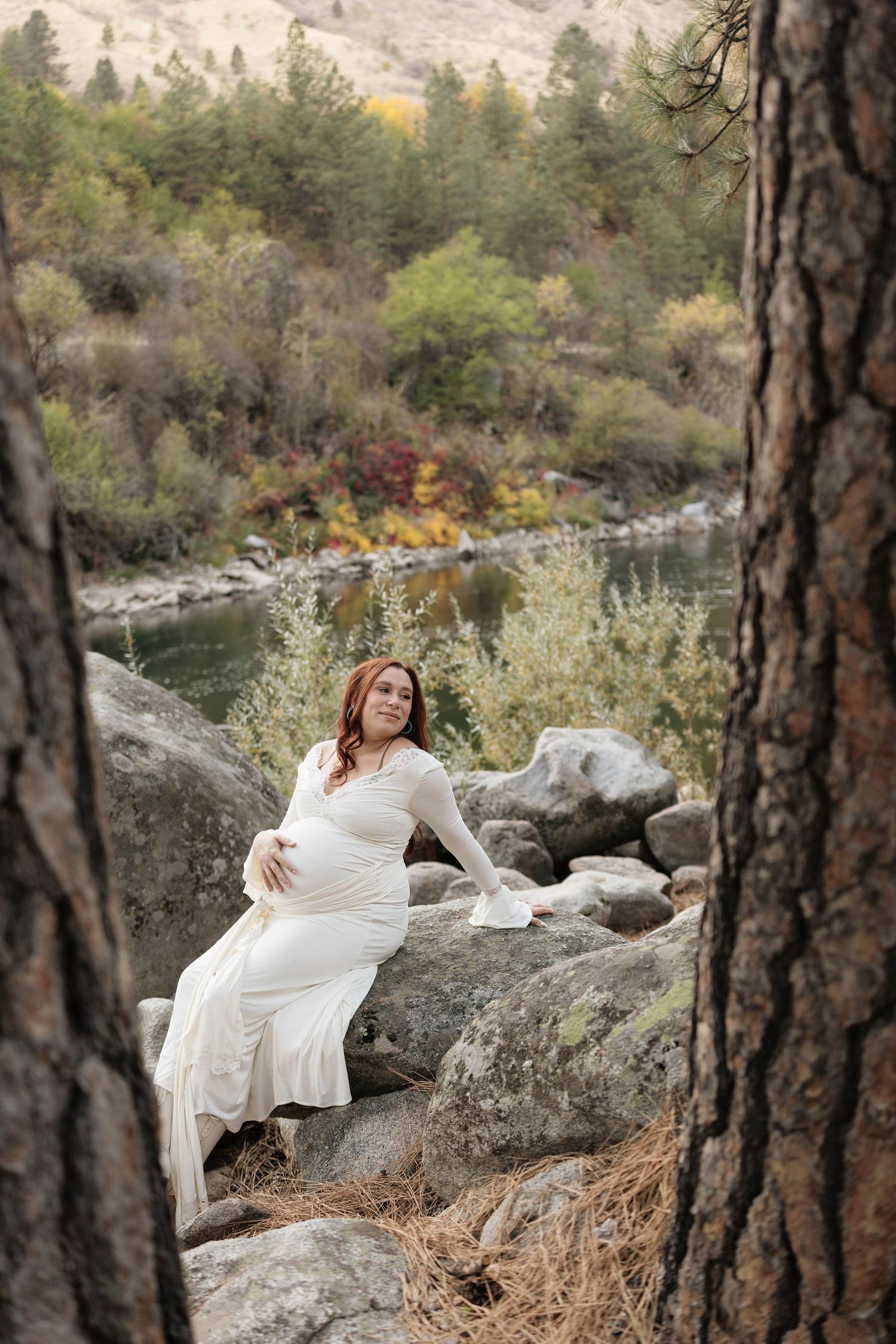 Pregnant woman in a flowing white maternity gown sits on a large boulder by a river, framed by pine trunks and autumn foliage.