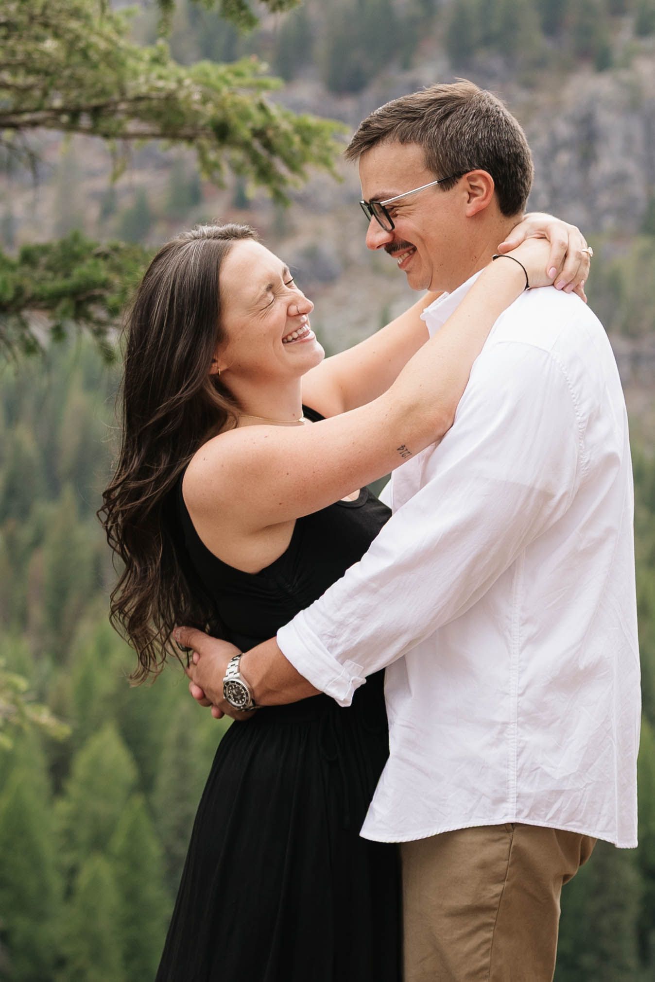 Smiling couple embracing outdoors, woman in black dress and man in white shirt, with a forested mountain valley in the background.