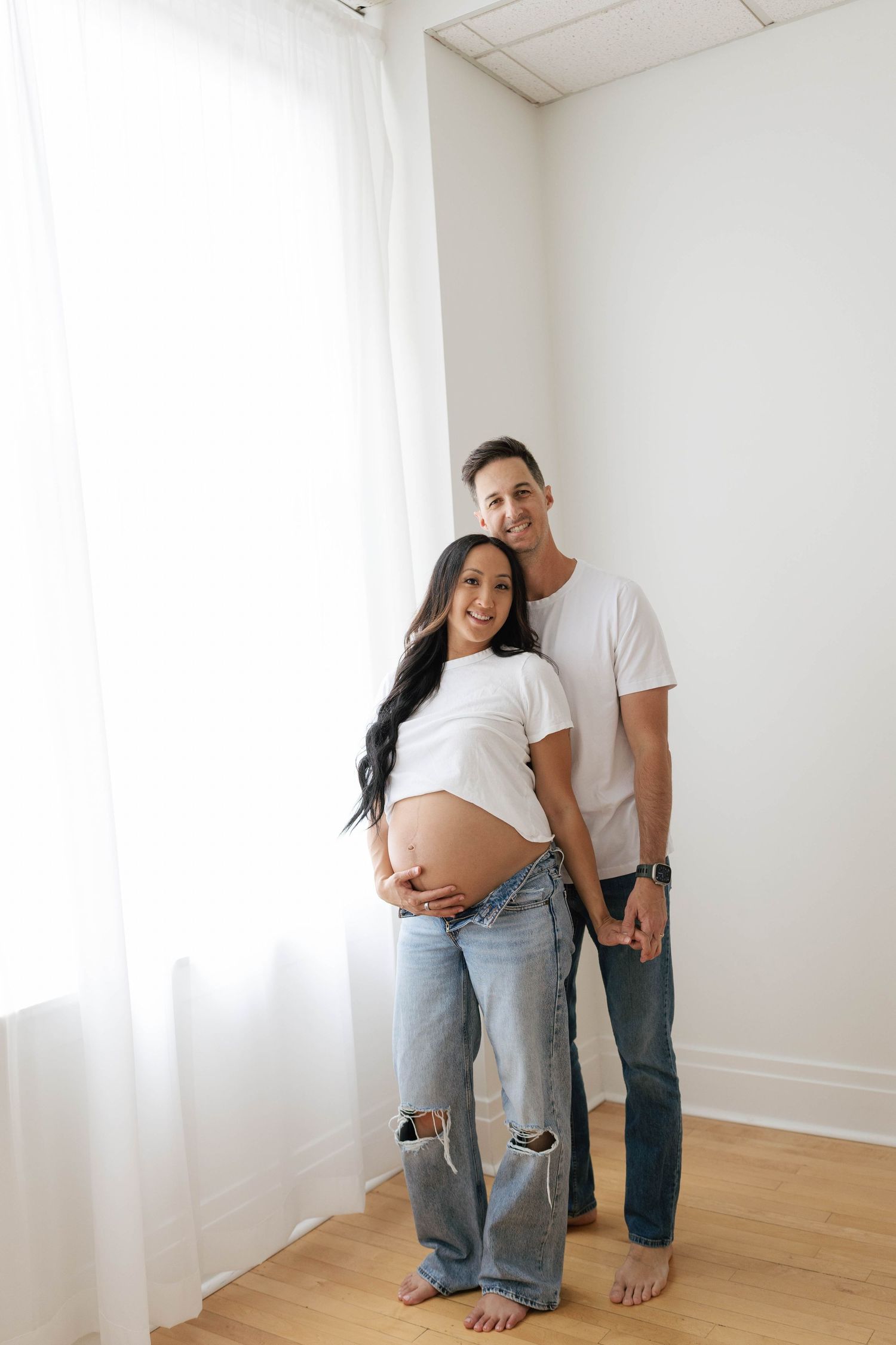 Smiling pregnant couple in white tees and jeans pose together in bright room, woman cradling baby bump.