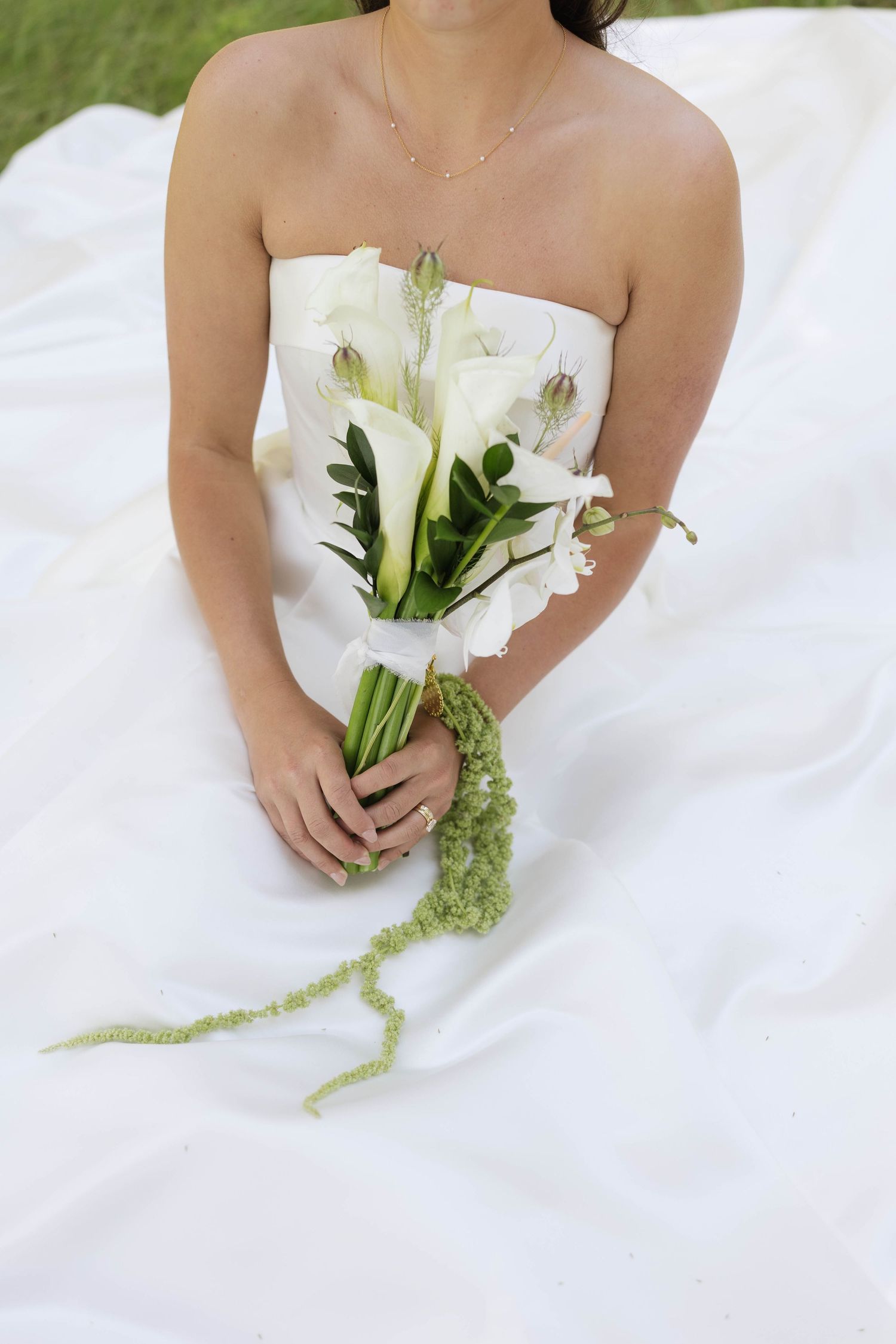 Bride in strapless white gown holding elegant calla lily bouquet with green trailing accents while seated outdoors.