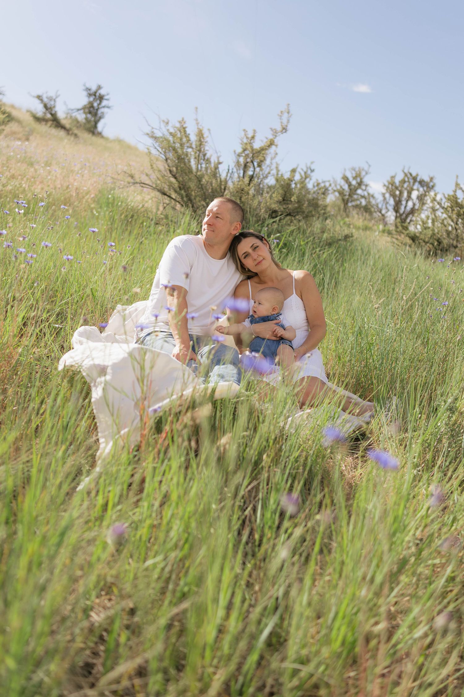 Couple sitting together in a lush green meadow with wildflowers, leaning on each other under a sunny blue sky.