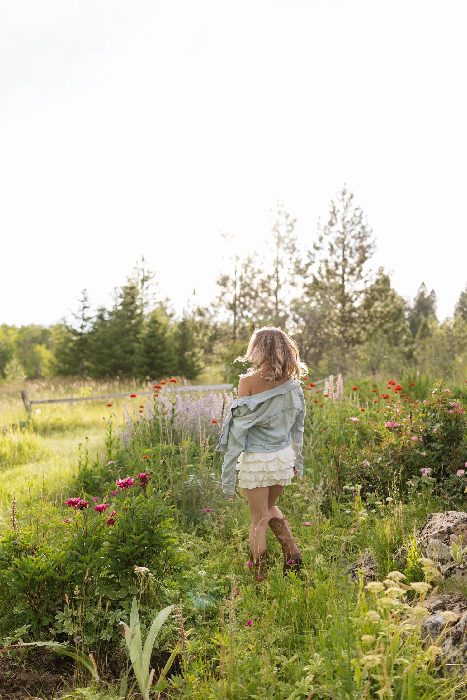 A young woman in a denim jacket and white skirt walks through a lush wildflower garden surrounded by tall trees.