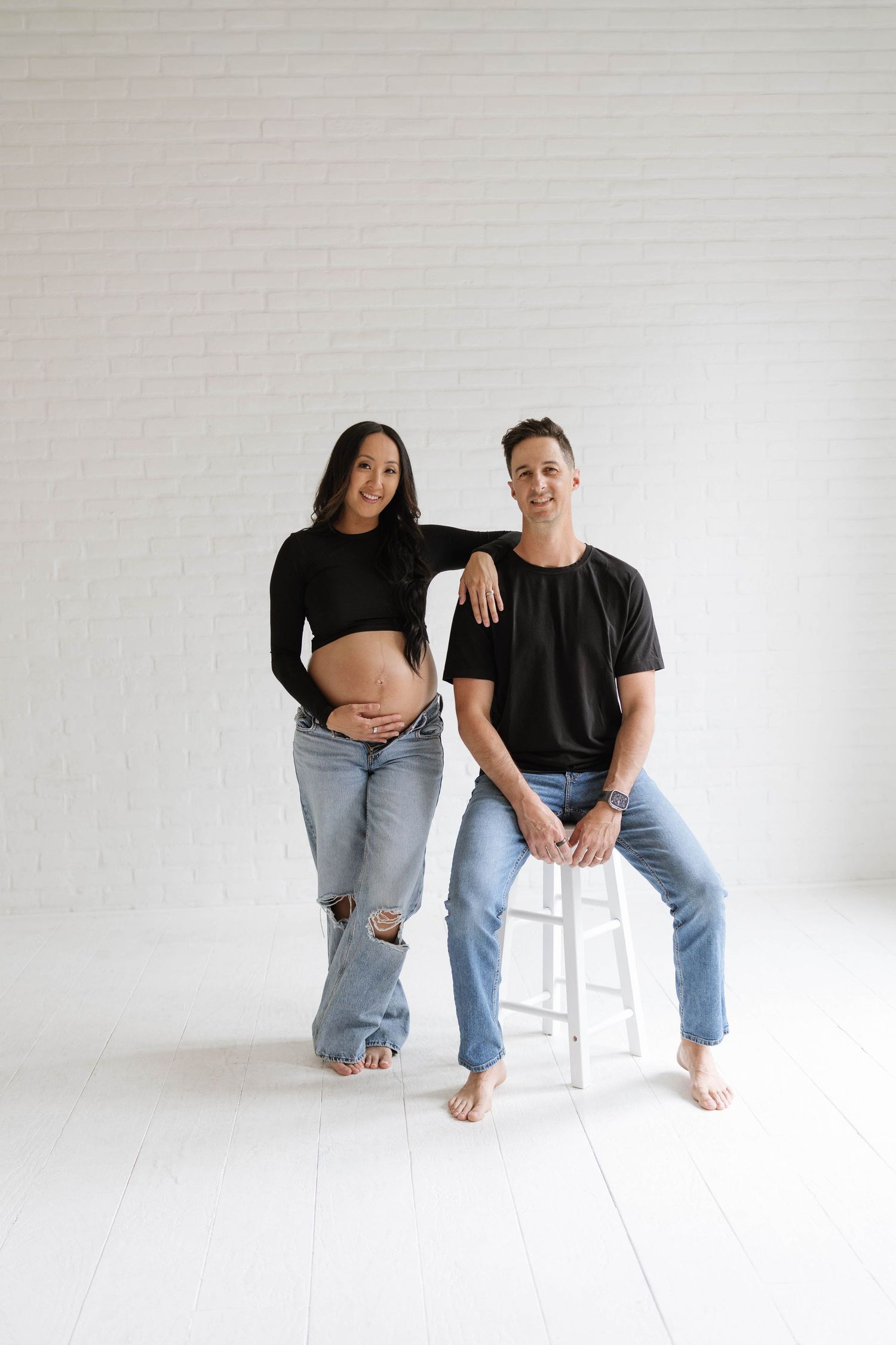 Pregnant couple in black tops and jeans posing in white studio, woman showing baby bump, man seated on stool.