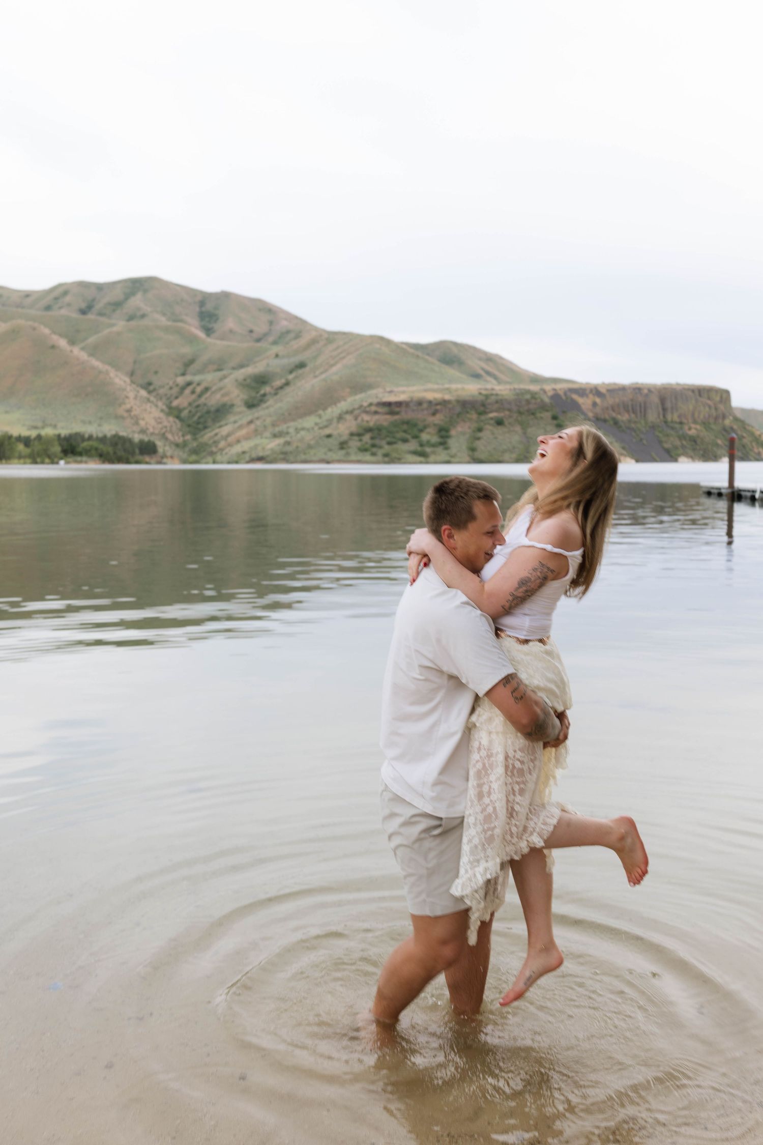 Couple laughing and embracing in shallow lake water with green hills in the background.