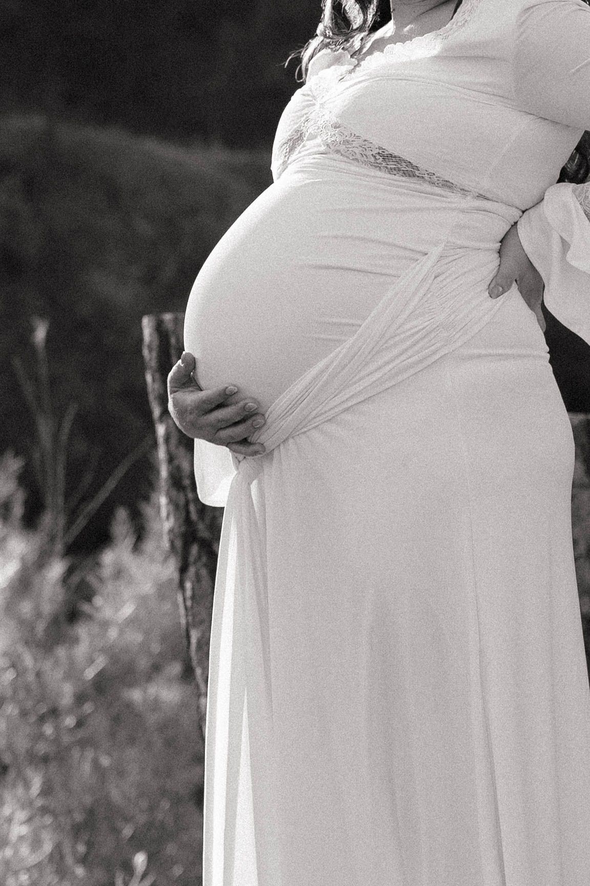 Black and white maternity photo of a pregnant woman in a flowing white gown cradling her baby bump outdoors near a wooden post.