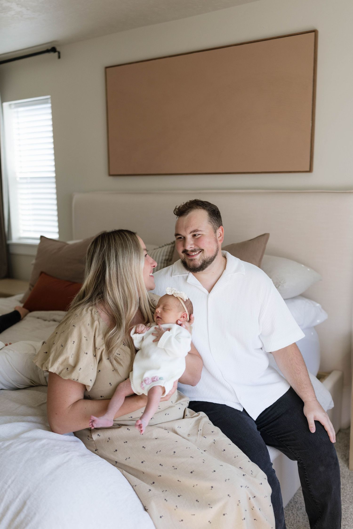 Smiling couple sitting on a bed in a bright bedroom, gently holding their sleeping newborn baby wrapped in a white outfit.