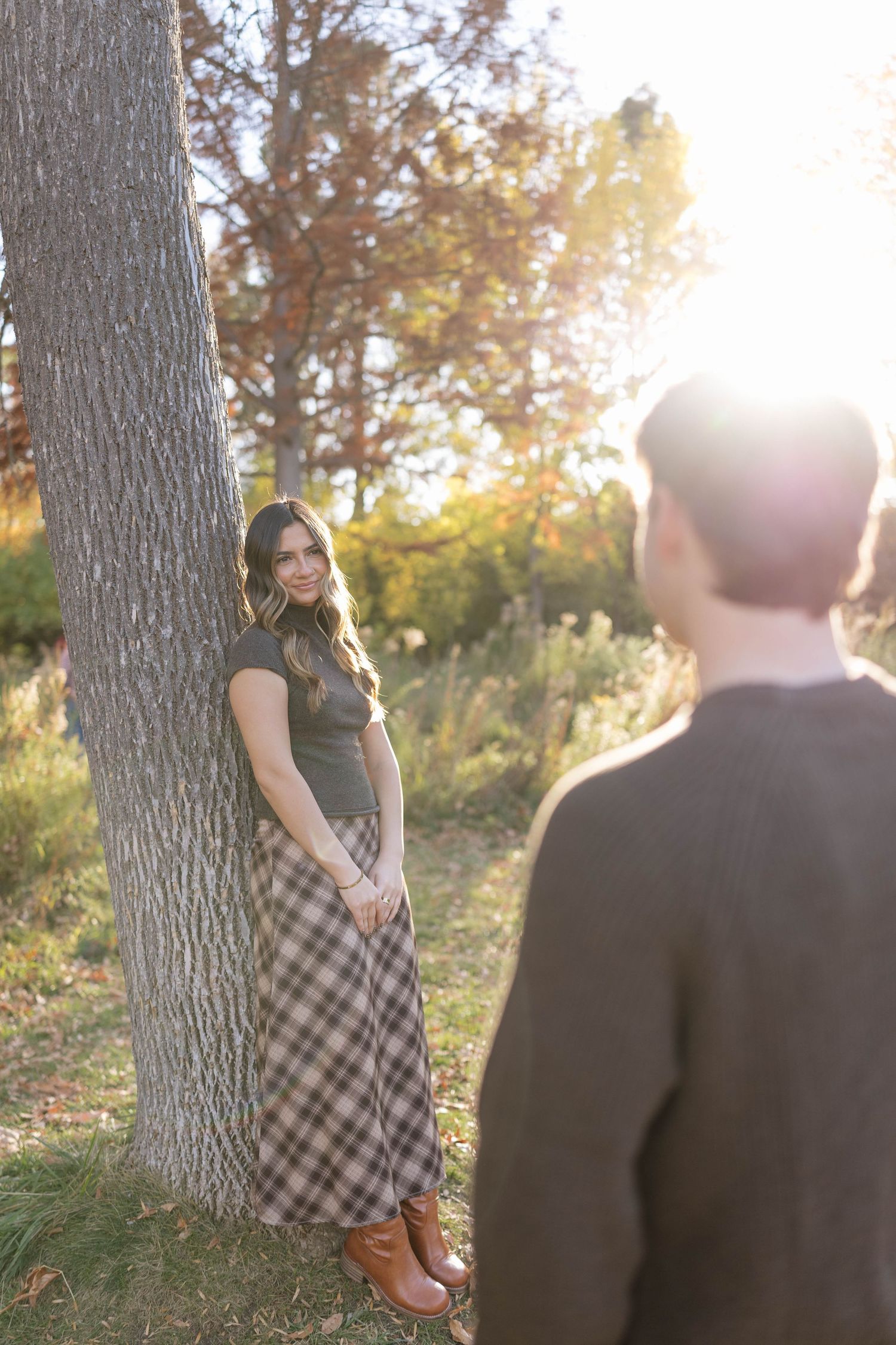 Woman in a plaid maxi skirt and brown boots leans against a tree in golden autumn sunlight, smiling at a person in the foreground.