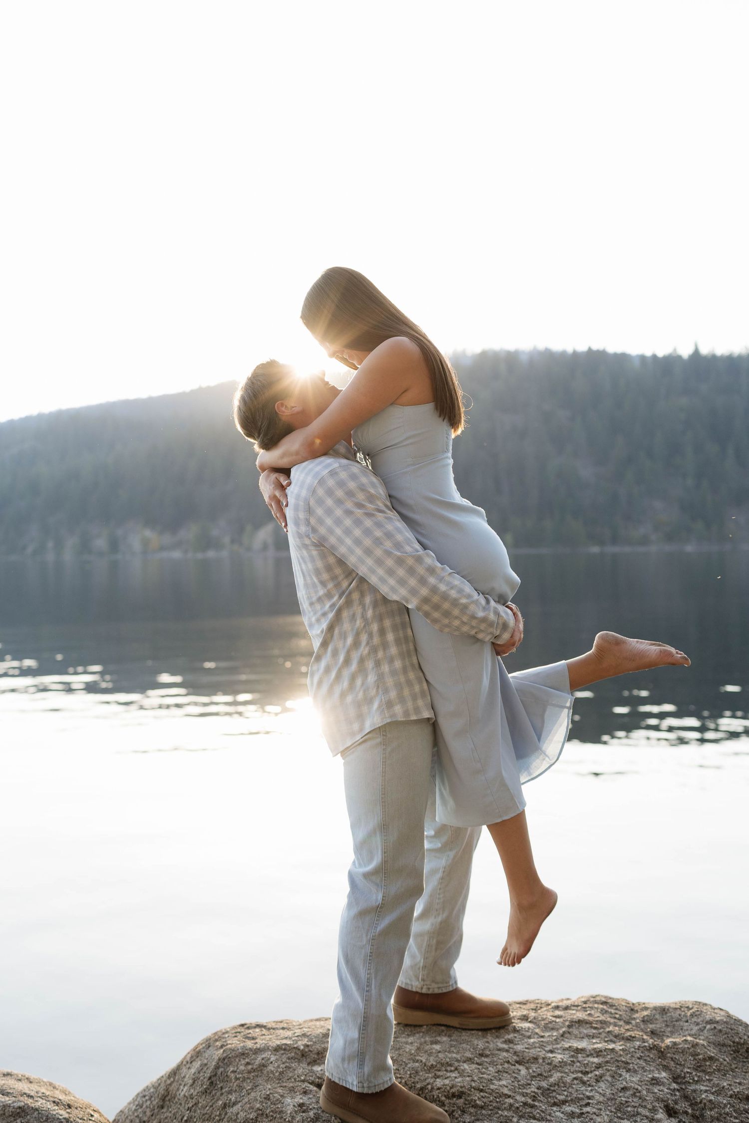 A romantic couple in matching gray and white outfits embraces by a serene lake during a sunset photoshoot.