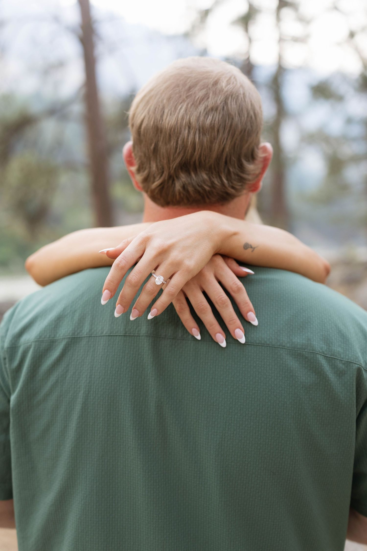 Woman's arms wrapped around a man's neck, showing her diamond engagement ring and manicured nails, with a blurred forest backdrop.