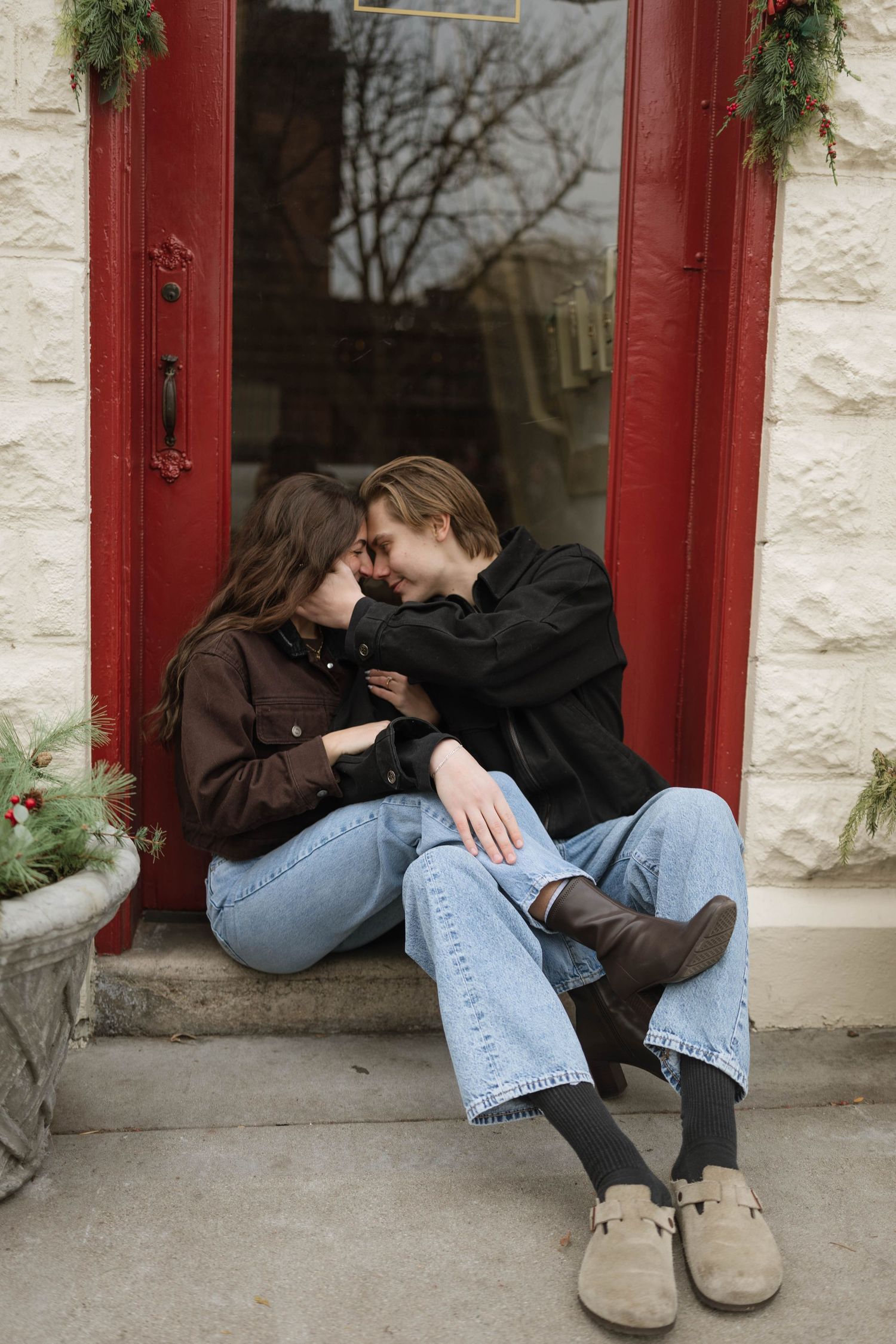 Two people in casual attire sit close together on a stone ledge against a red-trimmed building.