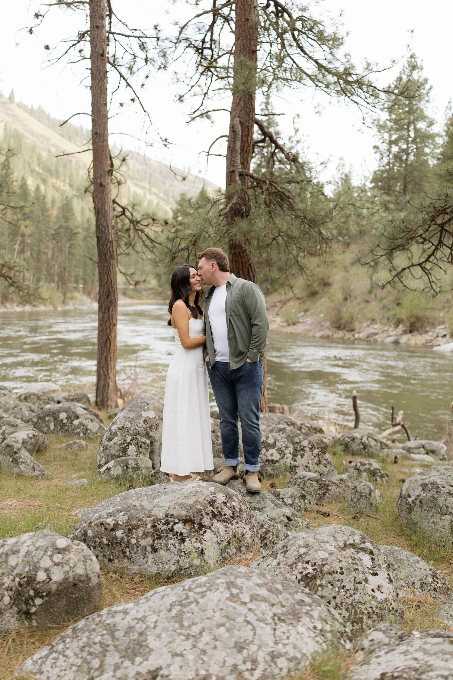Couple sharing a kiss on rocks by a river, surrounded by pine trees, woman in white dress and man in green jacket.