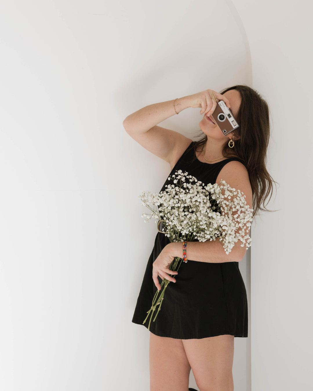Young woman in black outfit holding white baby's breath flowers, posing against a white wall with a camera.