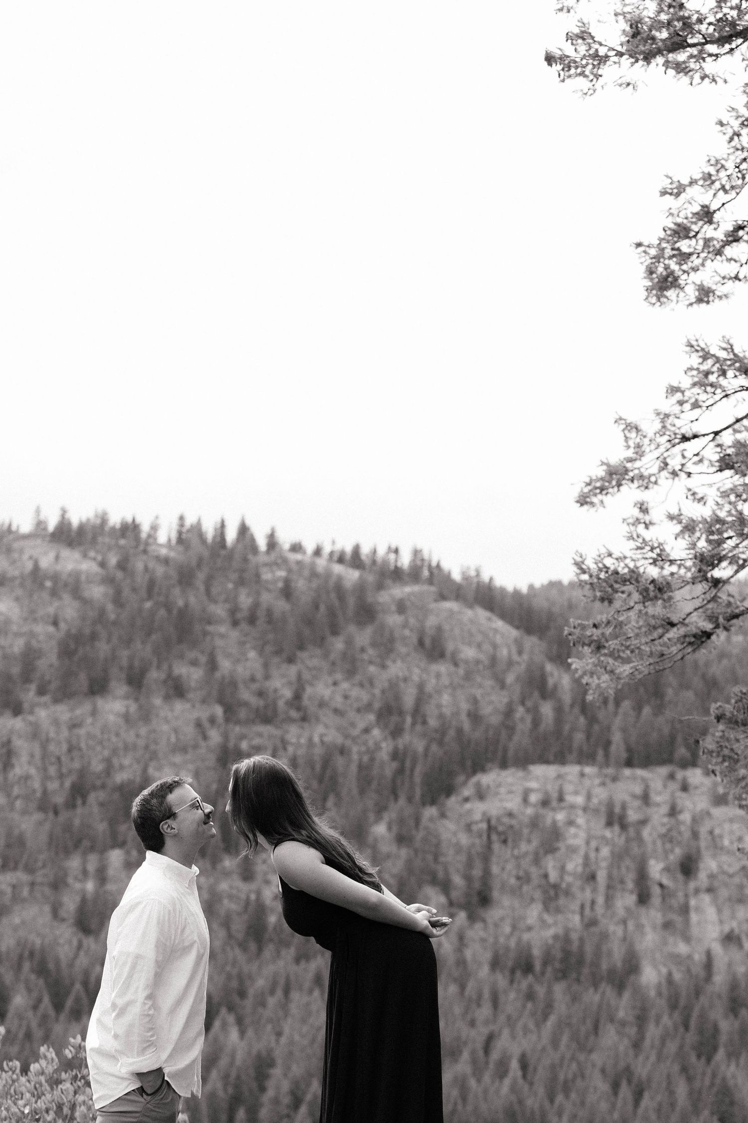 Black and white photo of a couple sharing a romantic moment outdoors with a forested mountain backdrop.